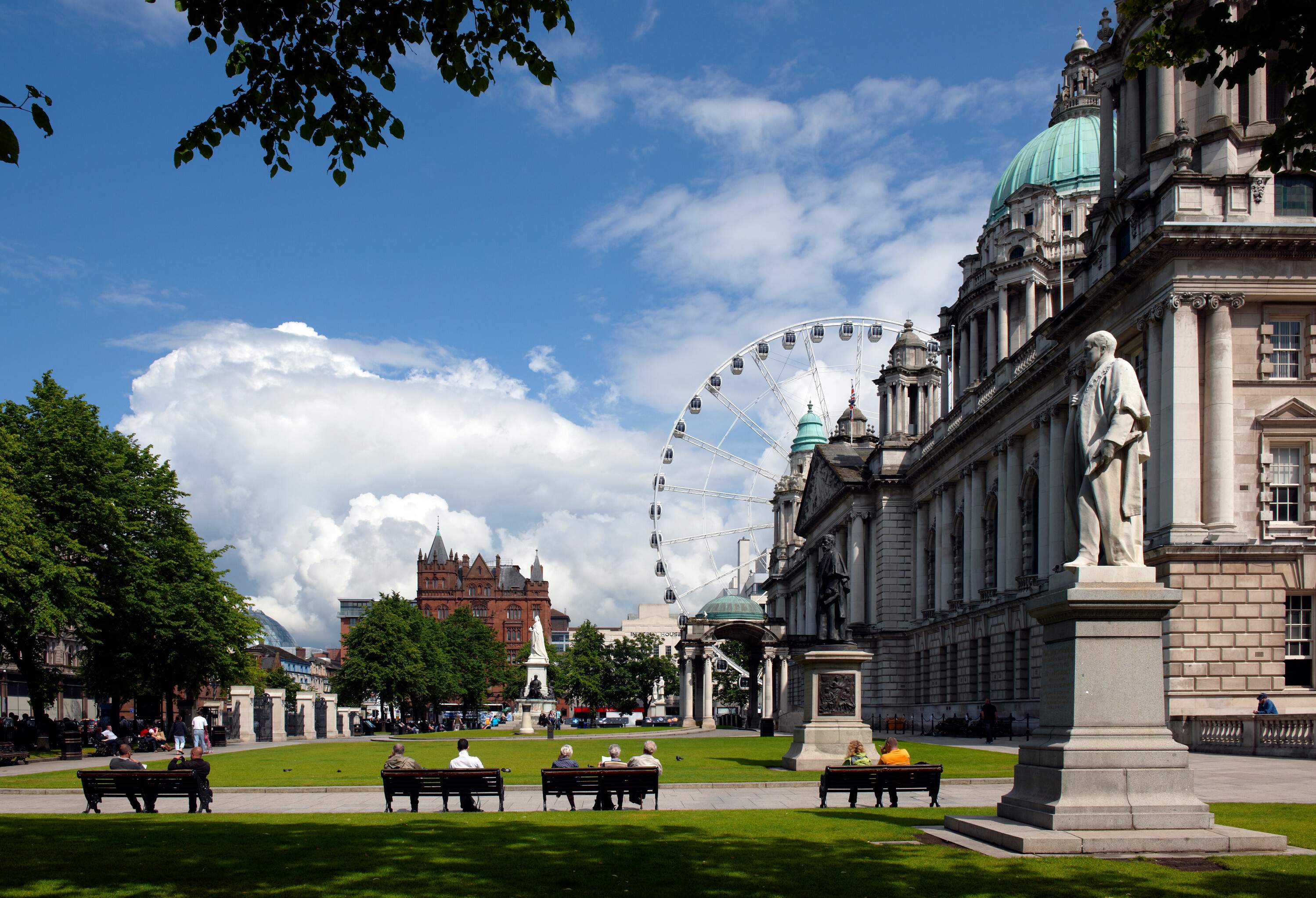 People sit on benches by the garden in front of a civic building with a view of an observation wheel against the cloudy blue sky.