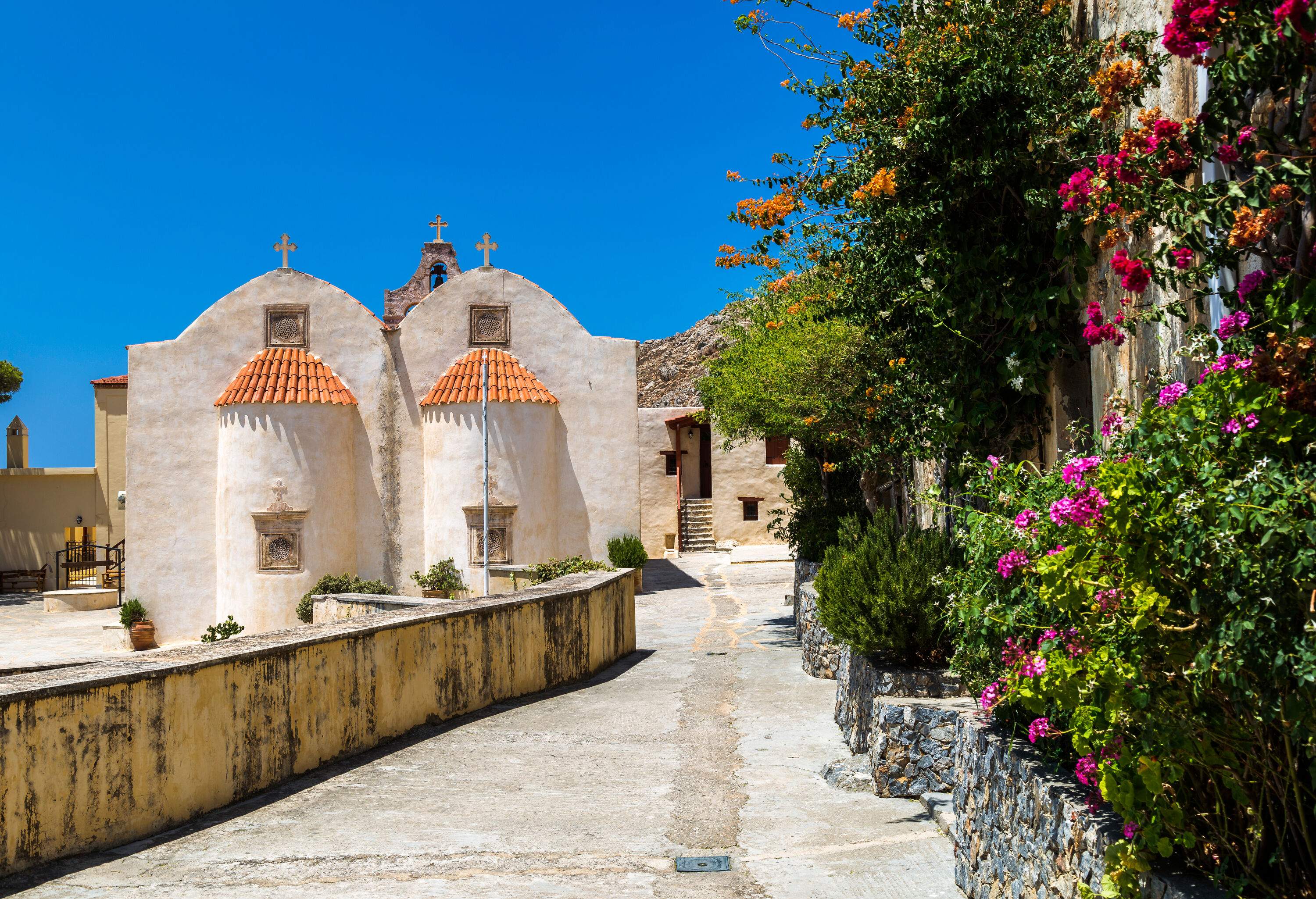 A concrete path along a lush garden towards a white monastery with red tile roofs and cross finials.