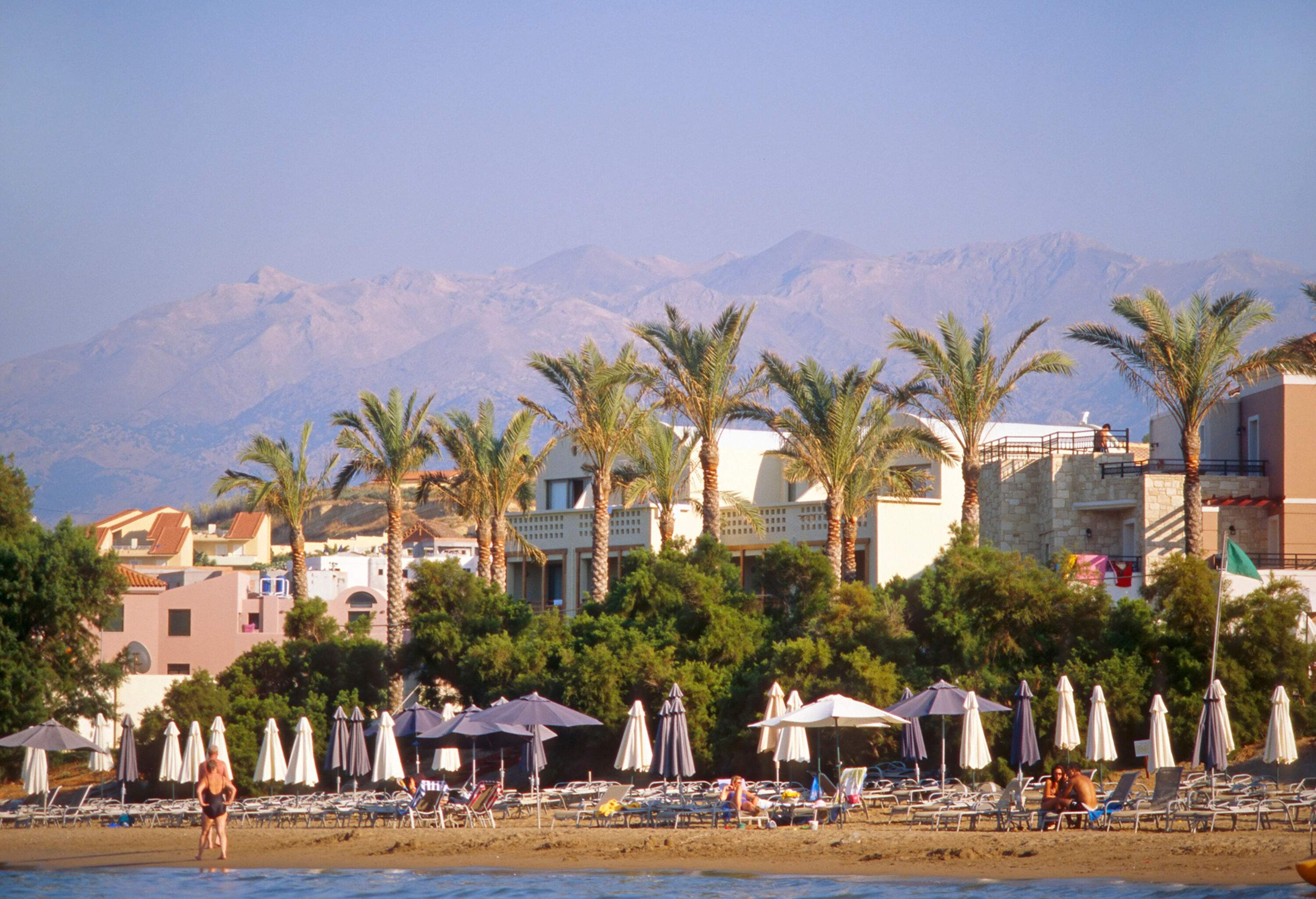 People relax on a beach with sunbeds and umbrellas along the waterfront buildings adorned with palm trees.