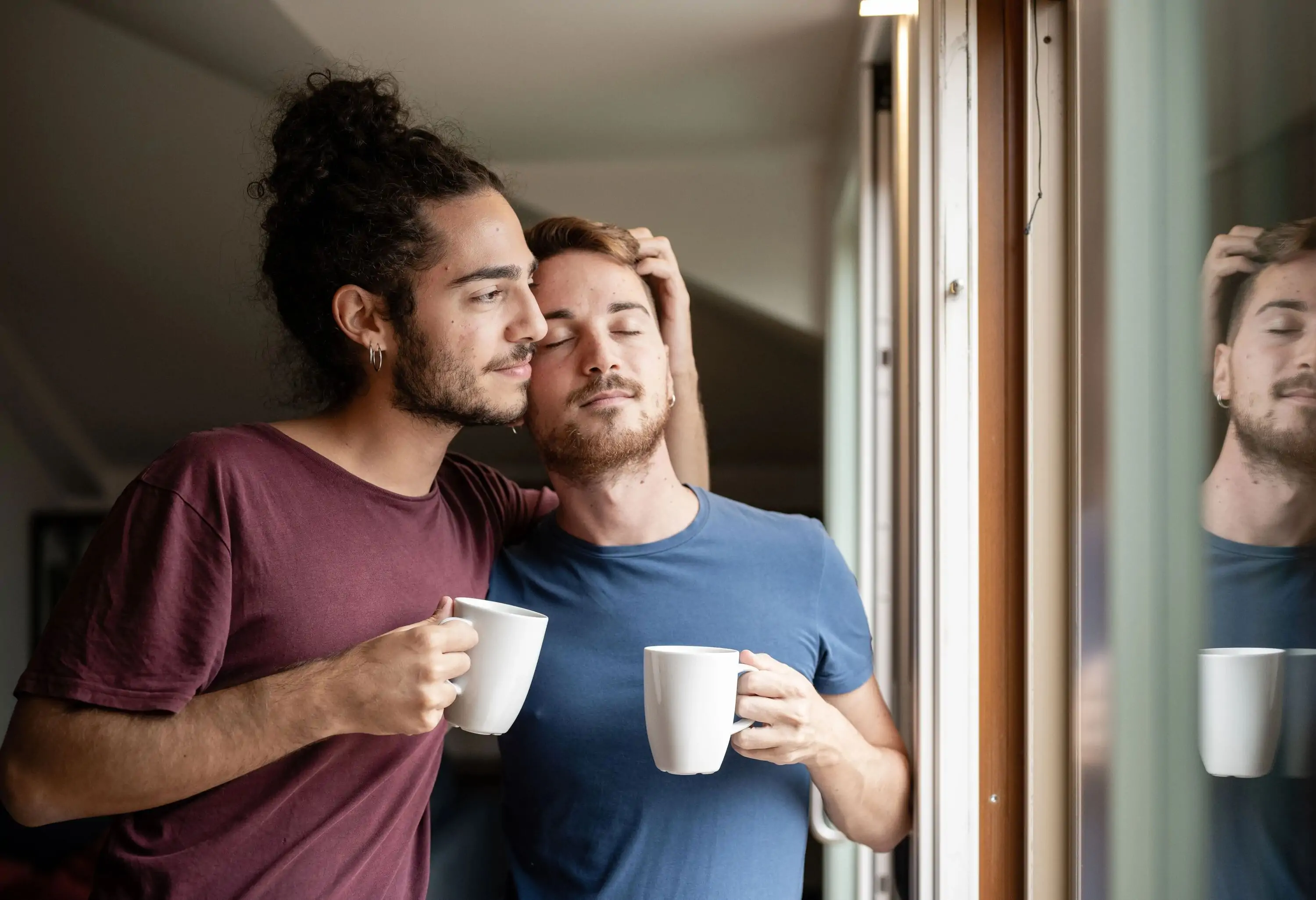 Gay couple sharing a special moment in the morning, holding cups of coffee by a window