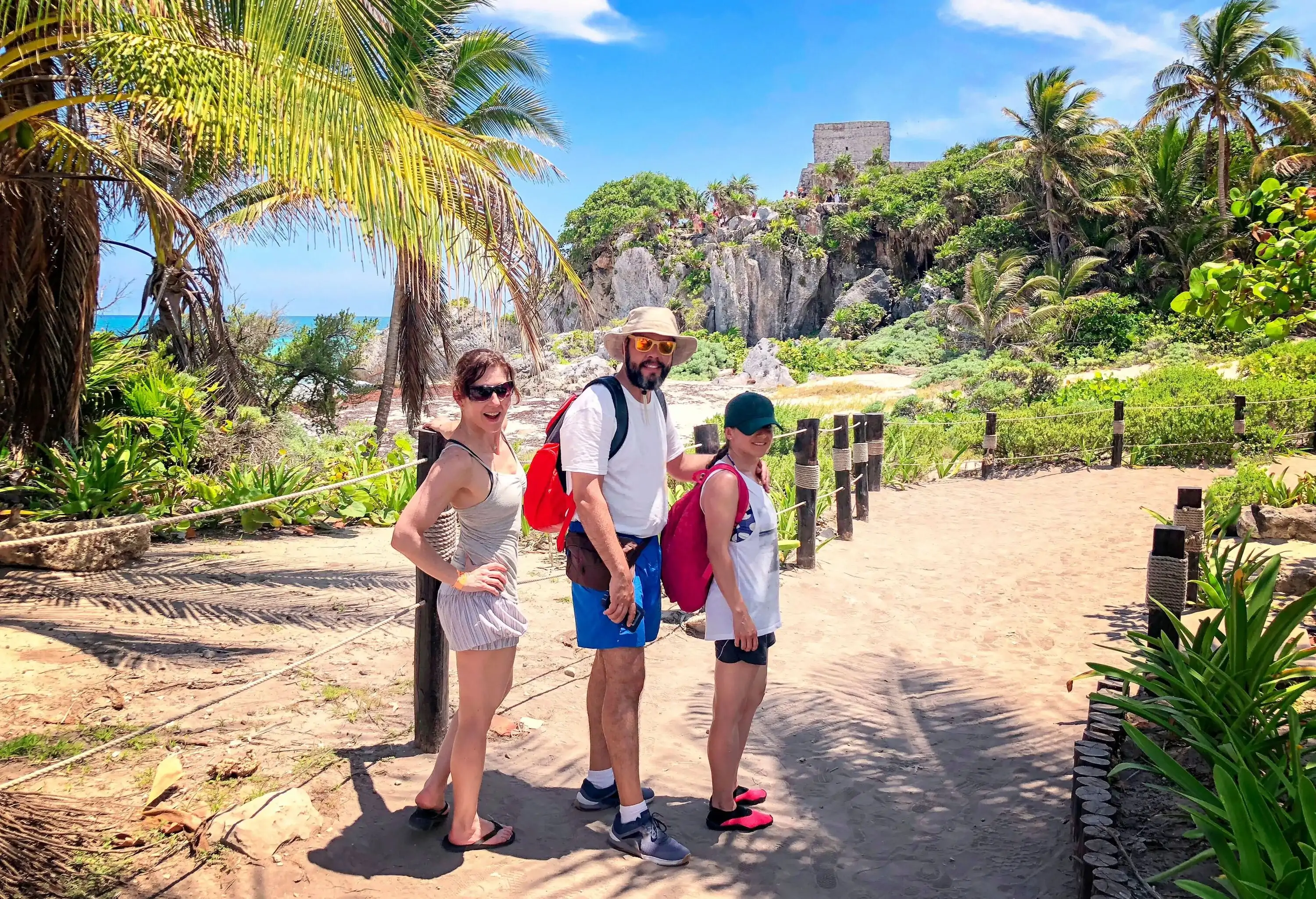 A radiant family of three, donning joyful smiles, walks along a sandy path, framed by lush tropical trees that provide a serene backdrop.