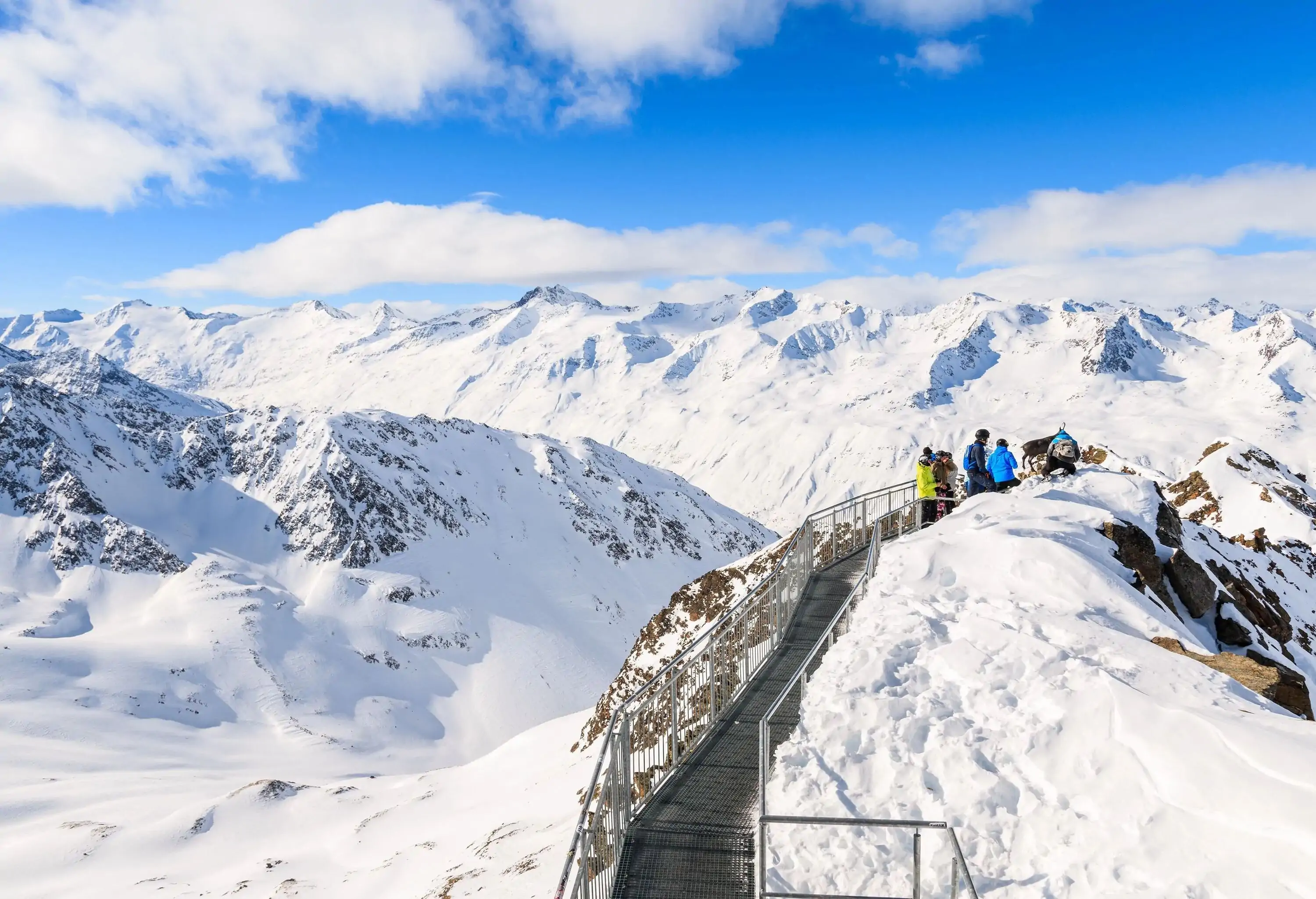 People standing on a mountaintop view deck looking out over a wide snow-covered mountain range.