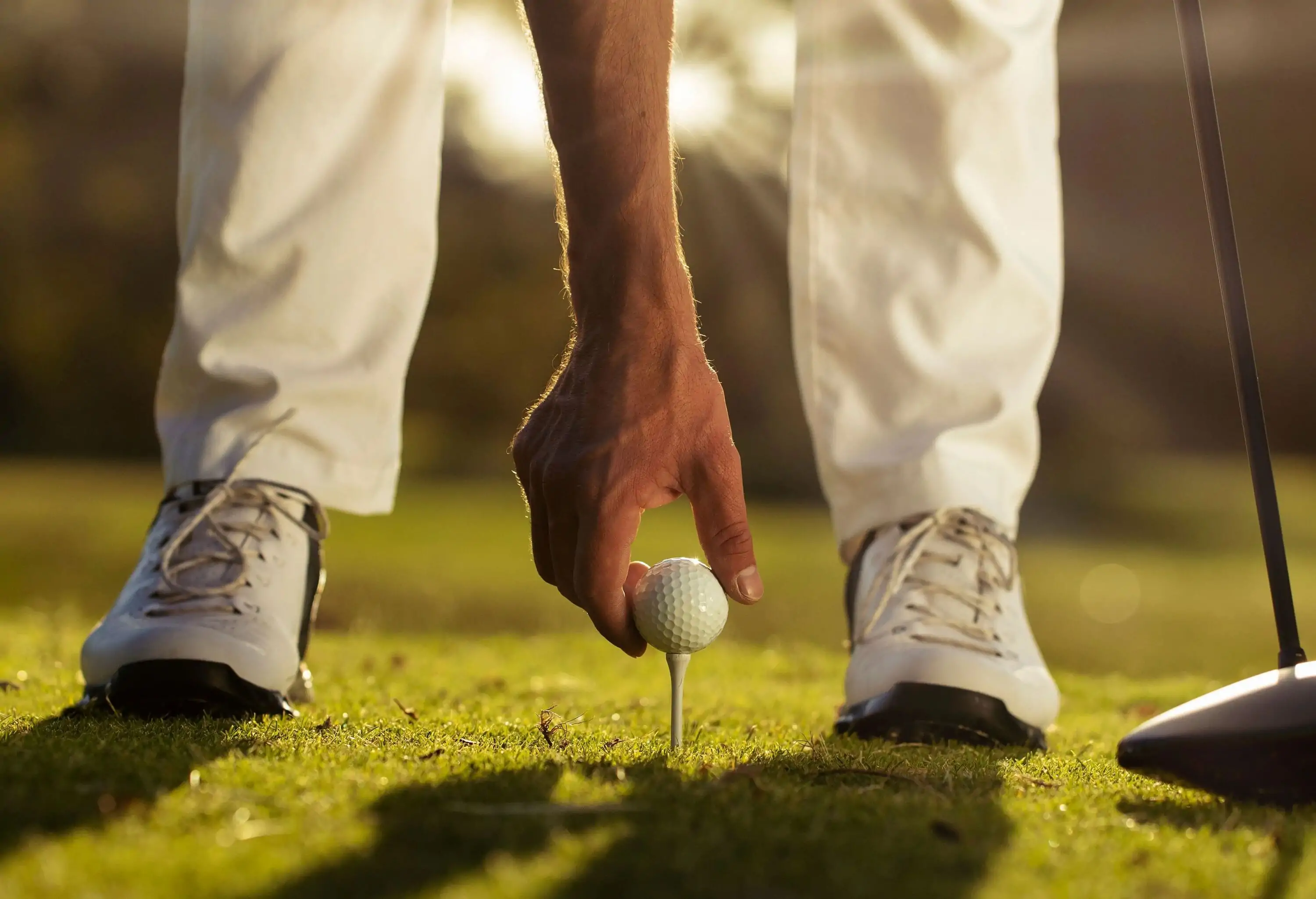 A golfer's hand puts the ball on top of the tee pin between his feet.