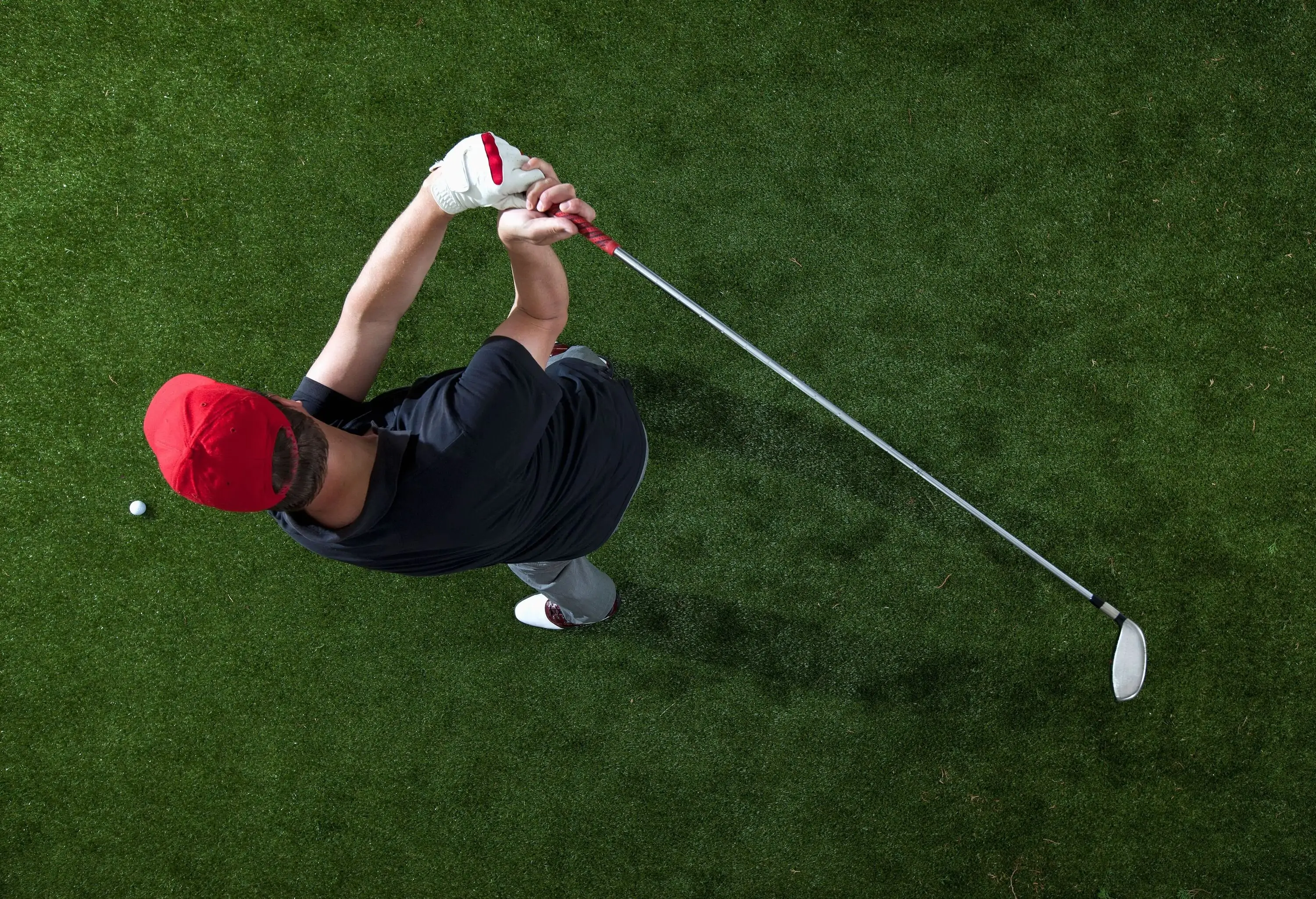 Top view of a person in a red hat on a golf course swinging a golf club.