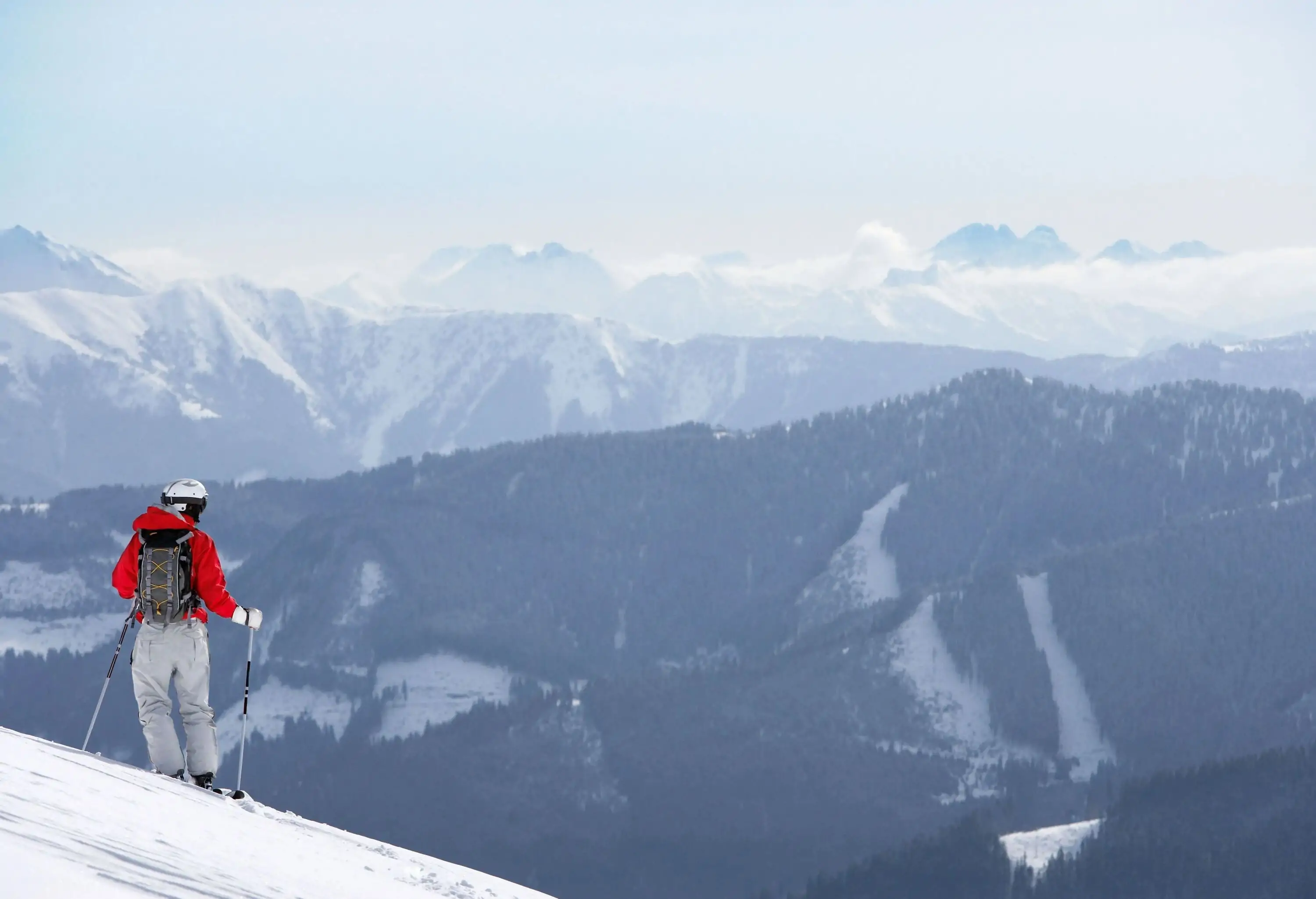 A skier in a red jacket standing on the slope side looking over the steep mountains.