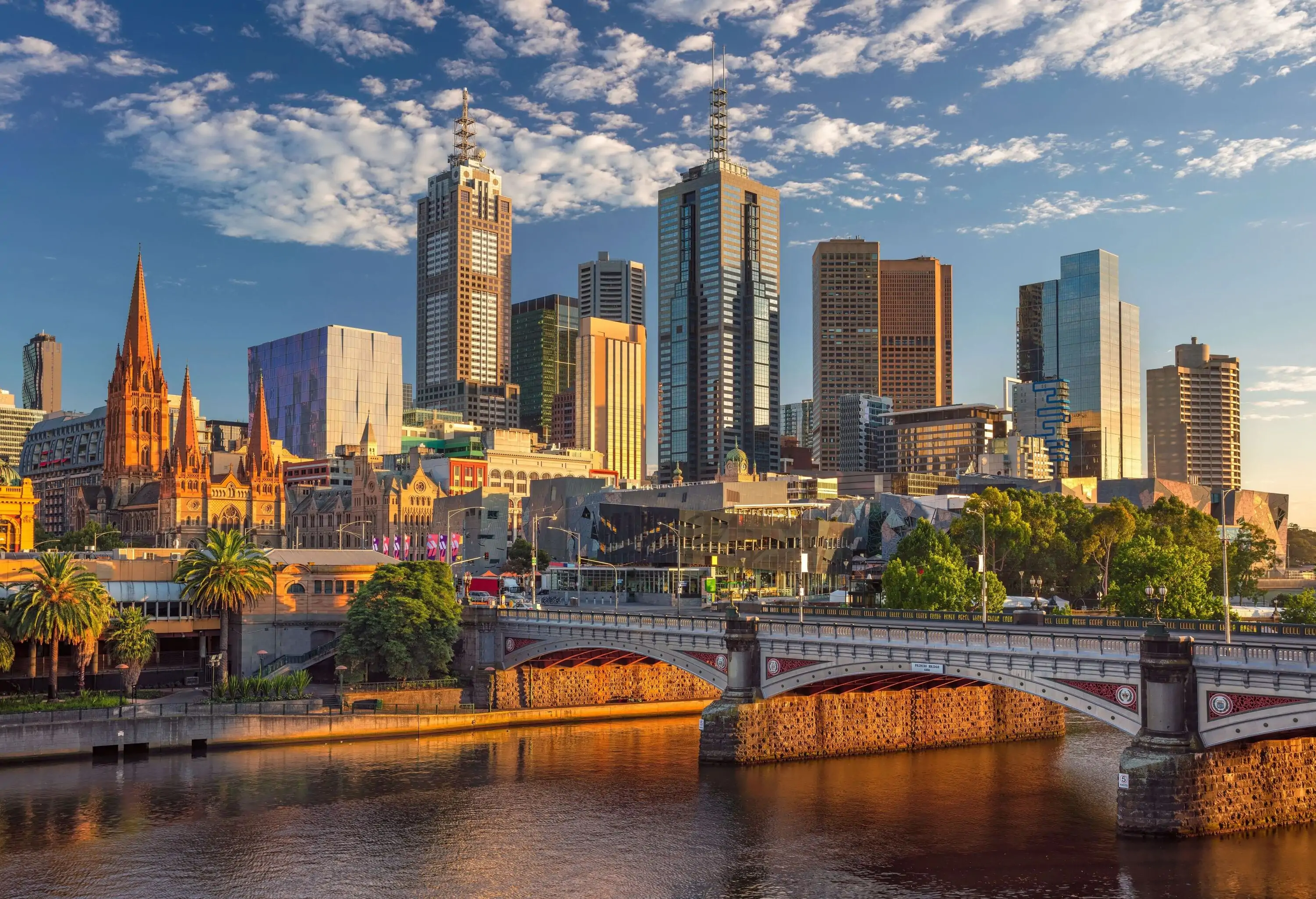 A contrasting skyline of historic spires and modern towering buildings on the bank of a river with an arch bridge.