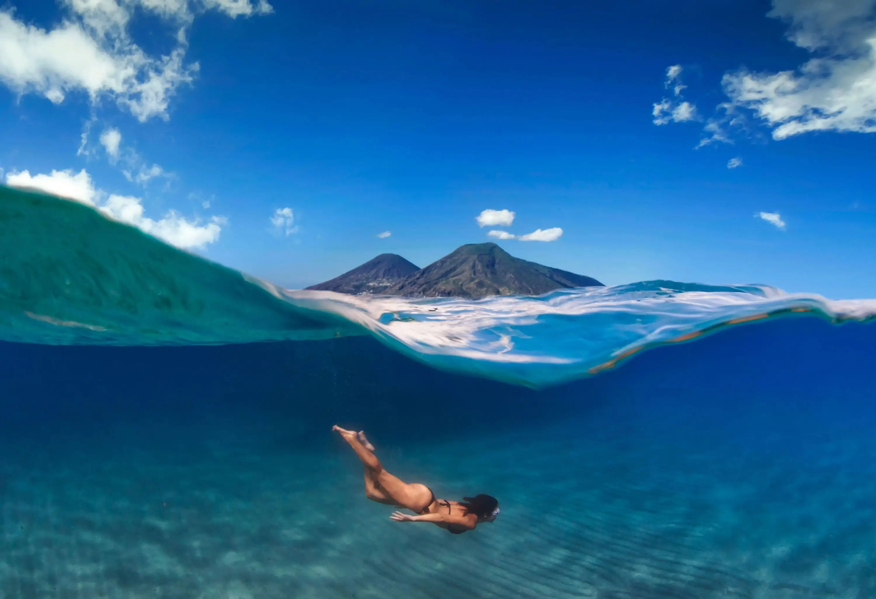 A woman diving deep into the sea, with mountains peeking over the wavy surface of the water.