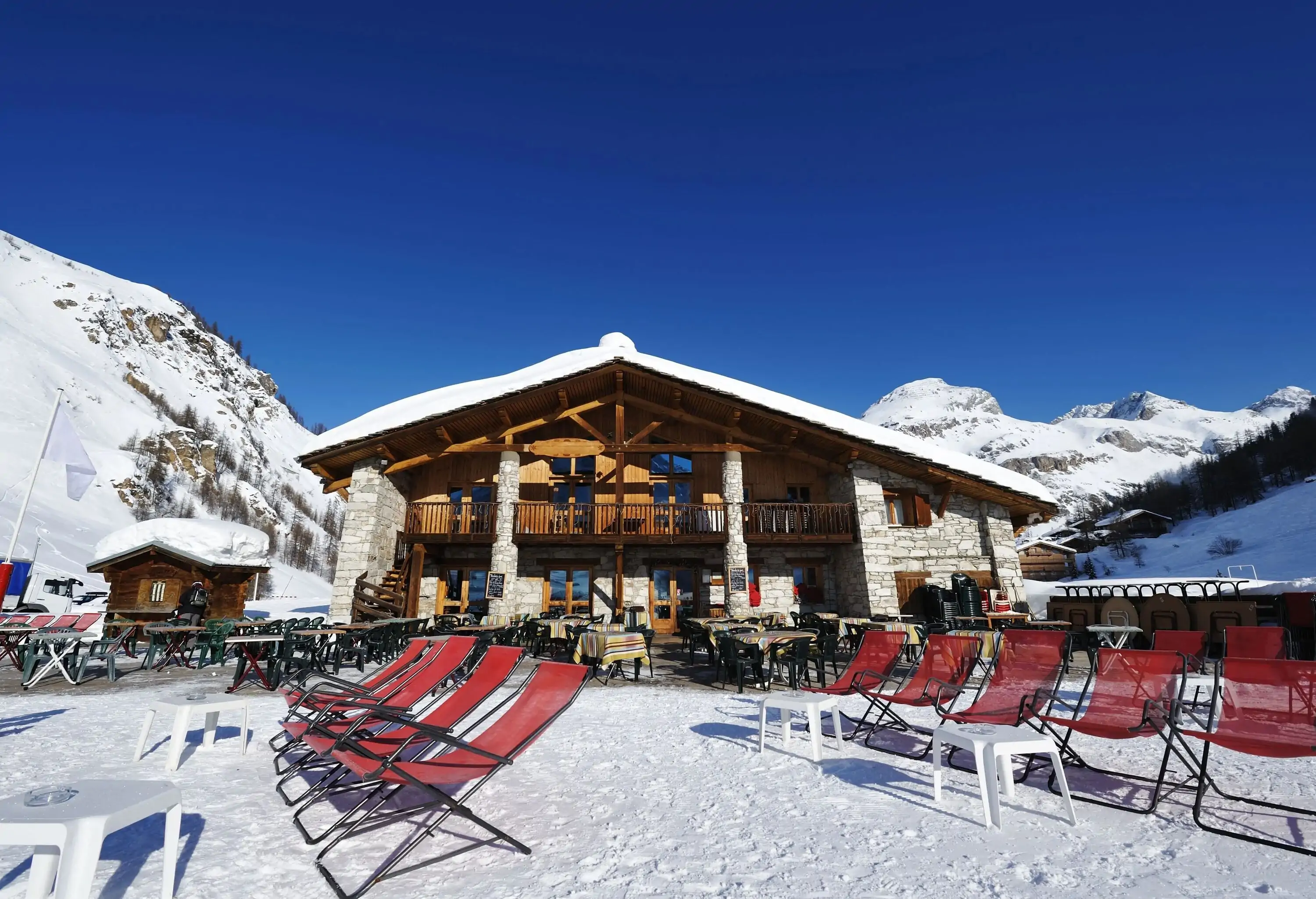 A row of folding chairs and an outdoor cafe in front of a large chalet, with a snowy mountain range in the background.