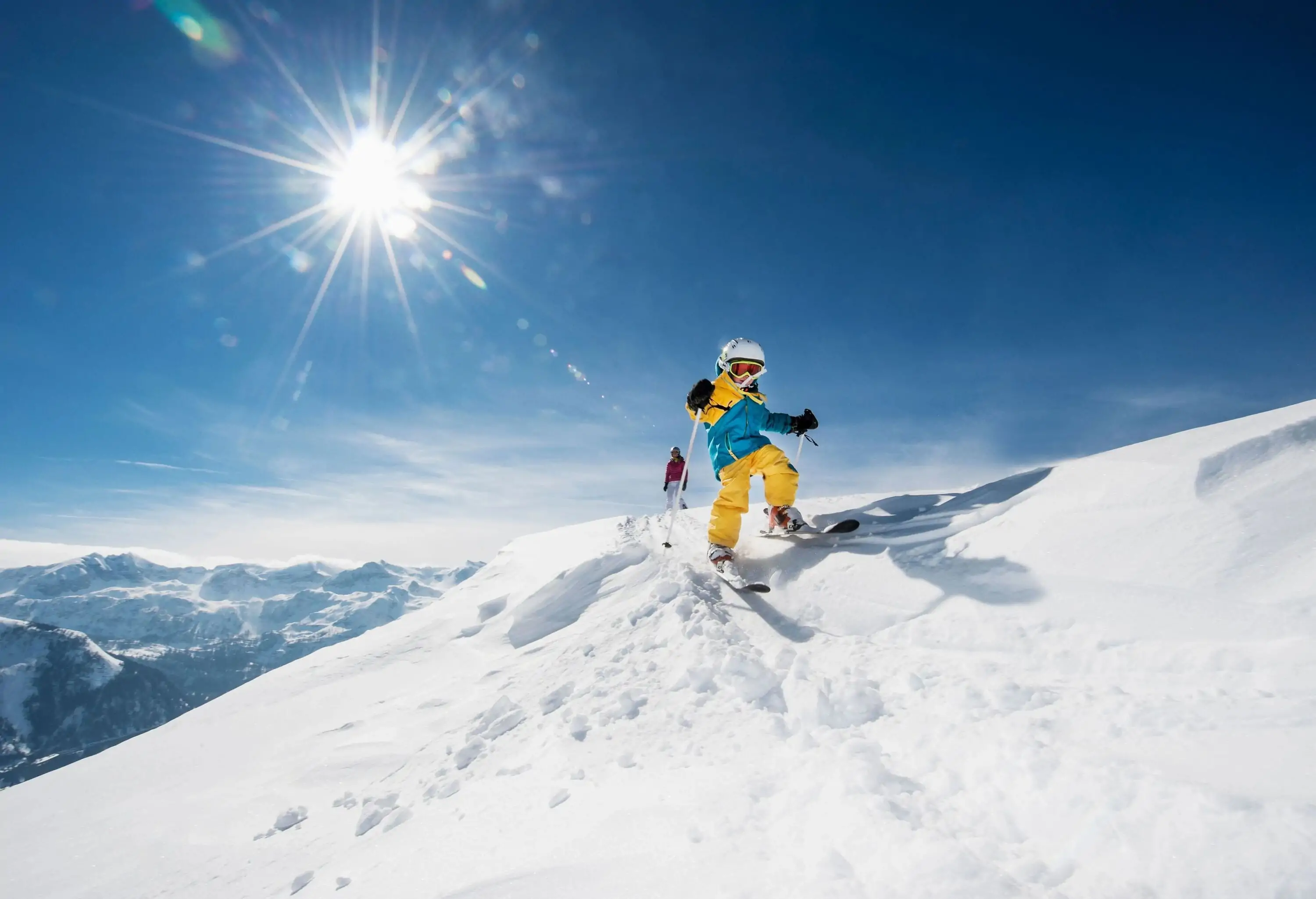 A skier wearing a yellow ski suit descends from the top of a mountain under the blue skies and bright sun.