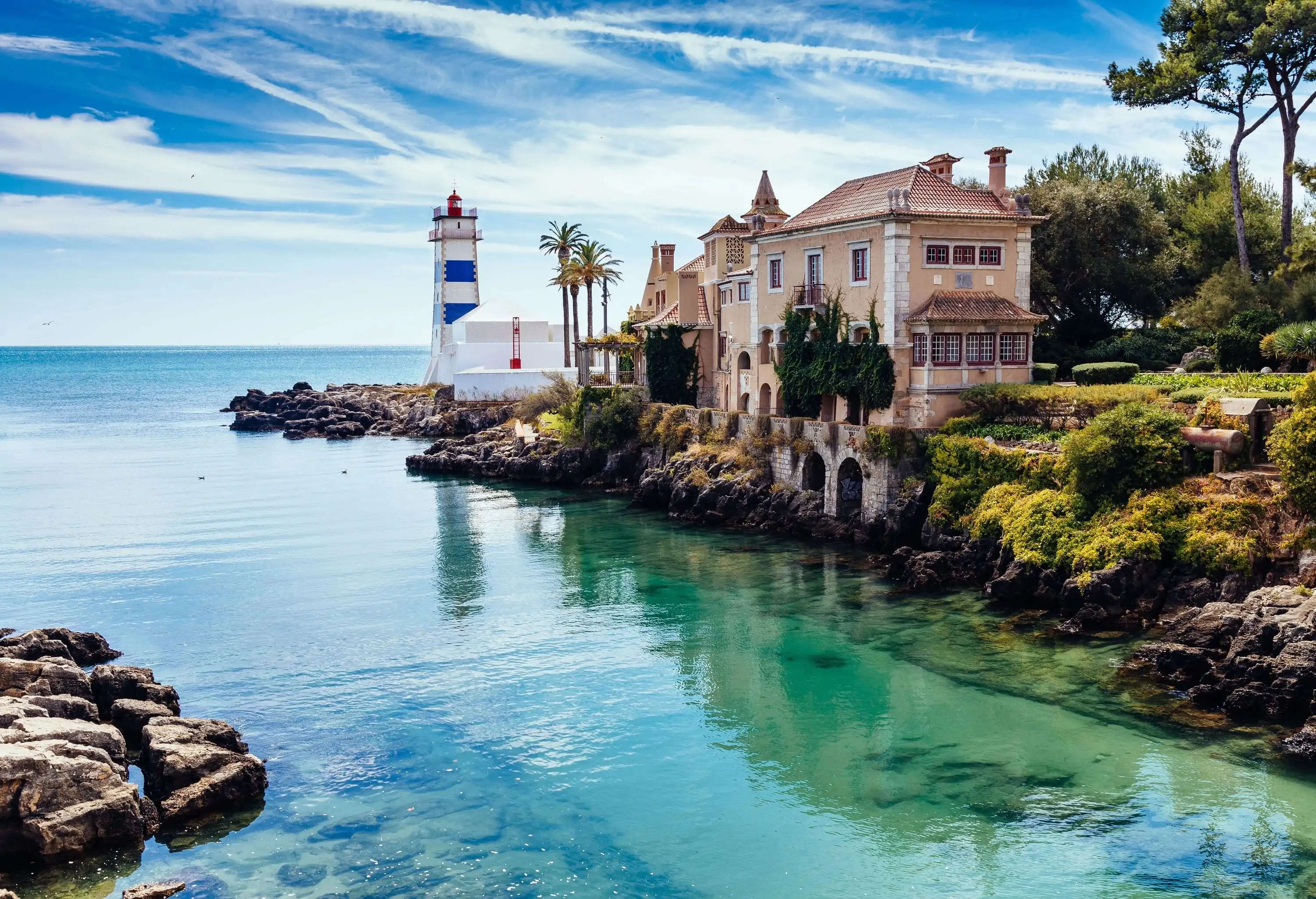 A blue and white striped lighthouse and a tiled-roof structure sitting on the rocky bank of a river.