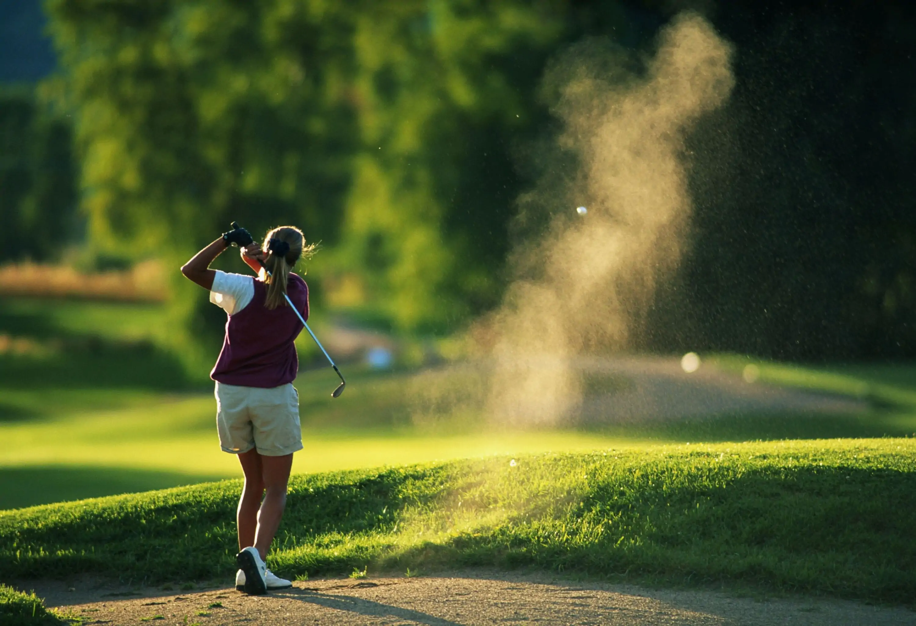 A female golfer hits a ball with a golf club with a spray of dust in a golf course.