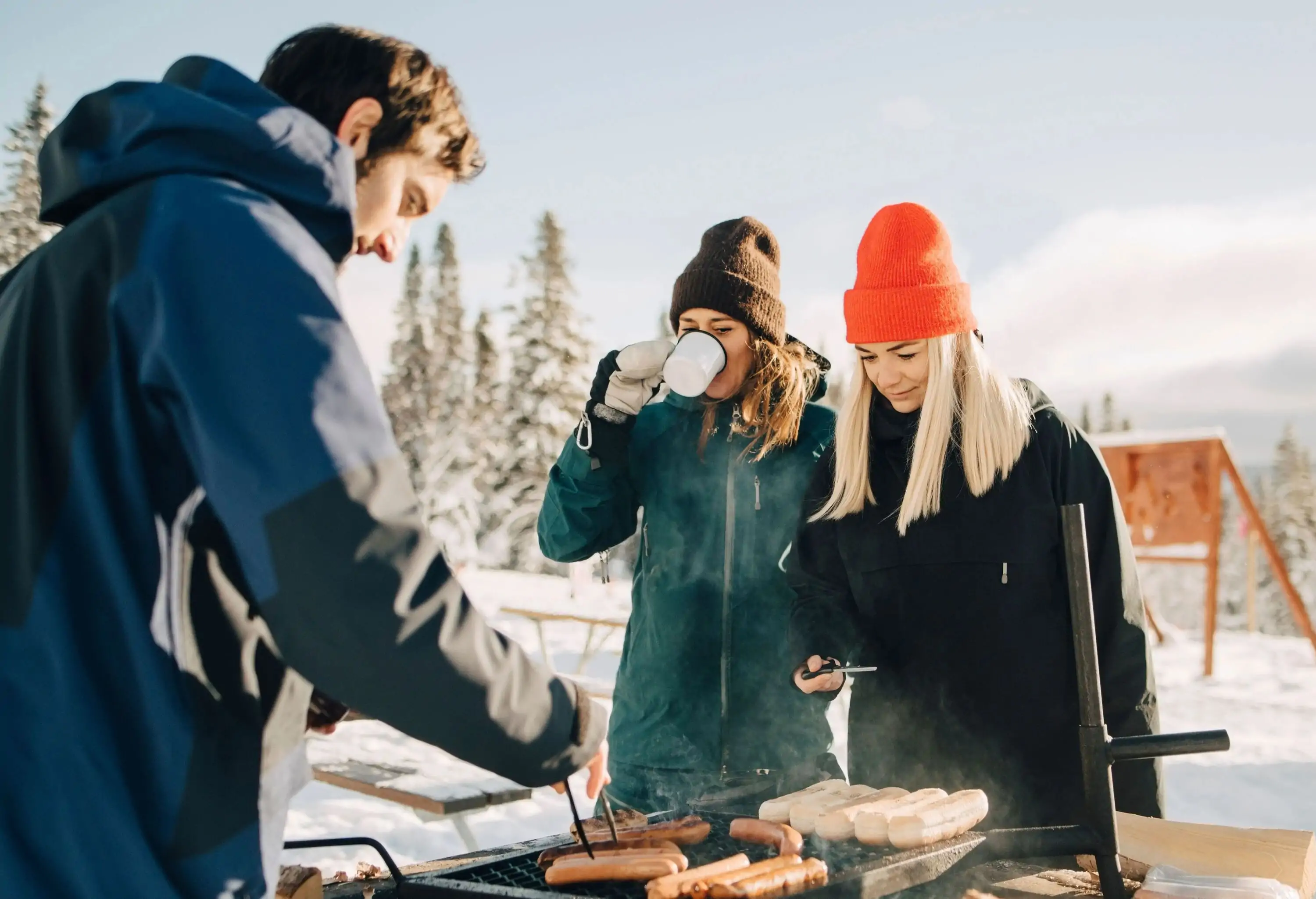 Three friends, one man and two women, grilling sausages in the snow on a sunny day