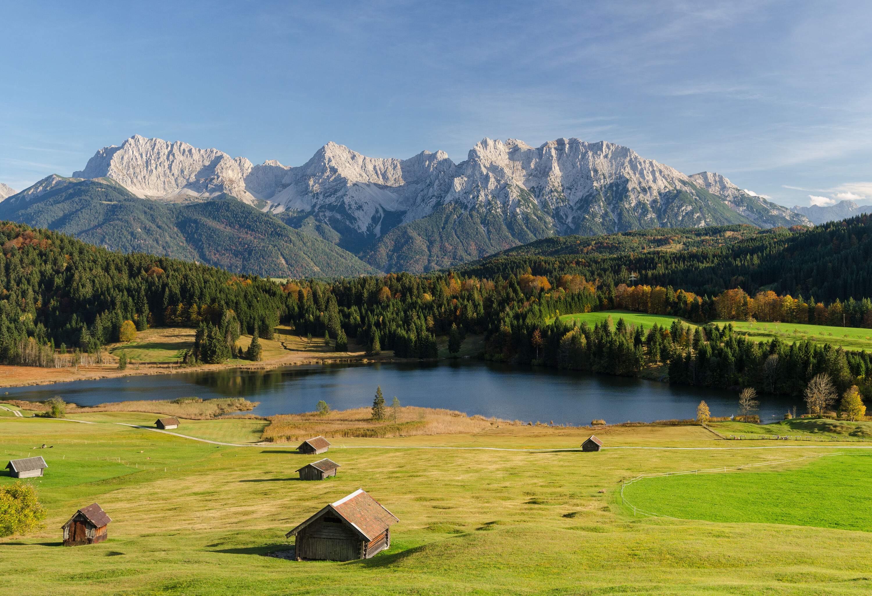 Haybarns in a green meadow by the lake surrounded by tall trees and a snow-capped mountain range in the background.