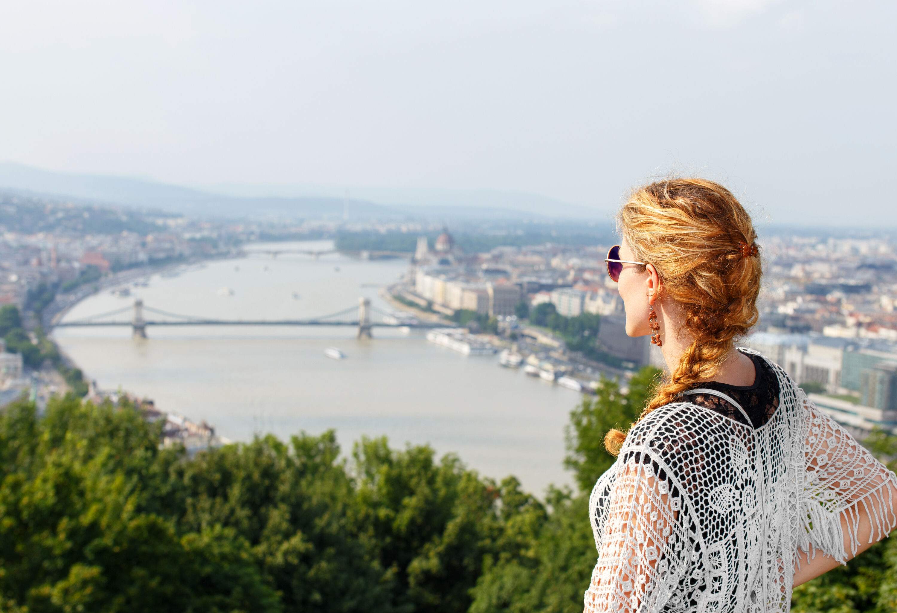 A young woman wearing sunglasses stands on high ground with a view of the bridge across a broad river in the middle of a crowded city.