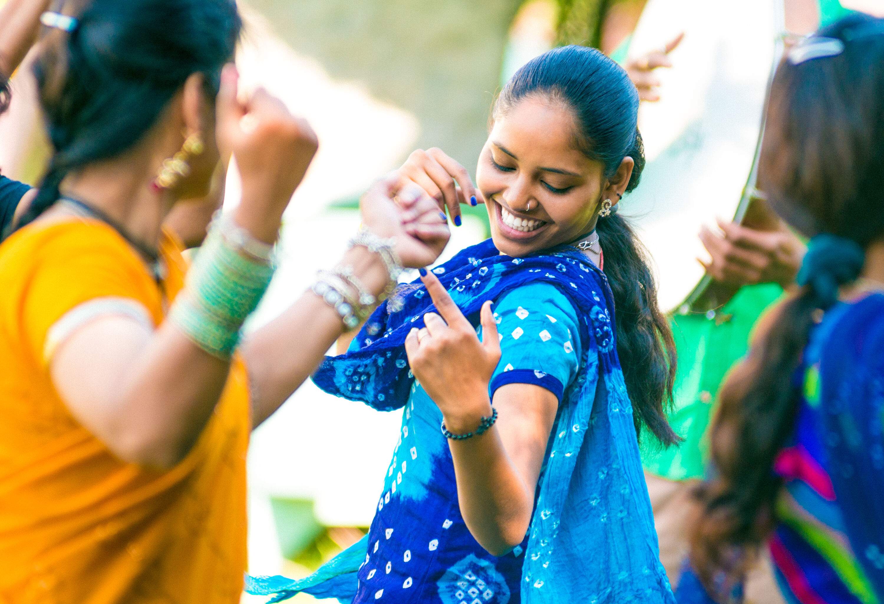 A young Indian woman twirls and sways with her friends, her vibrant and colourful dress adding to the joyous atmosphere as they dance together in celebration.