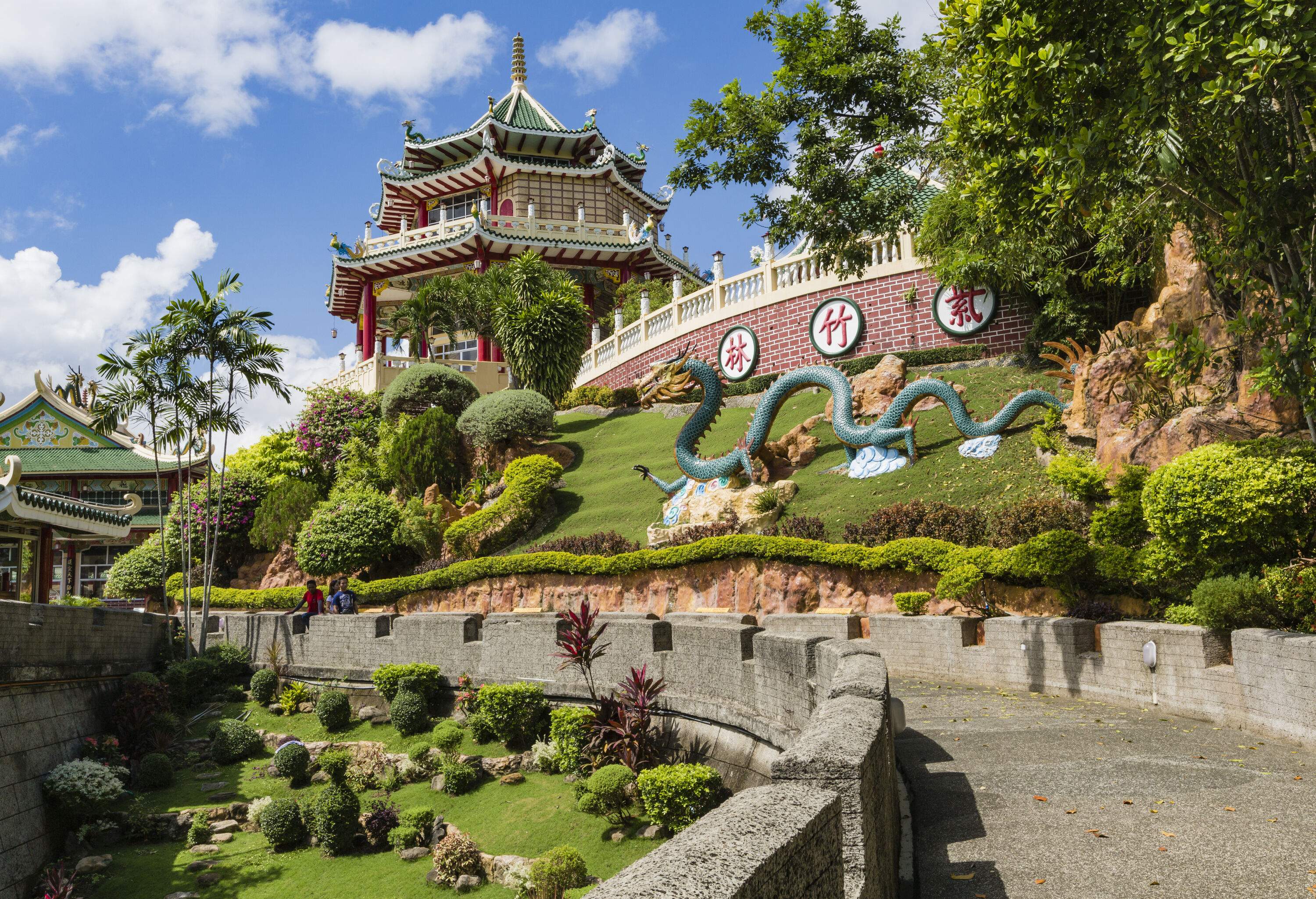 A statue of a dragon decorated in a lush park of a colourful multi-tiered temple on a hilltop.