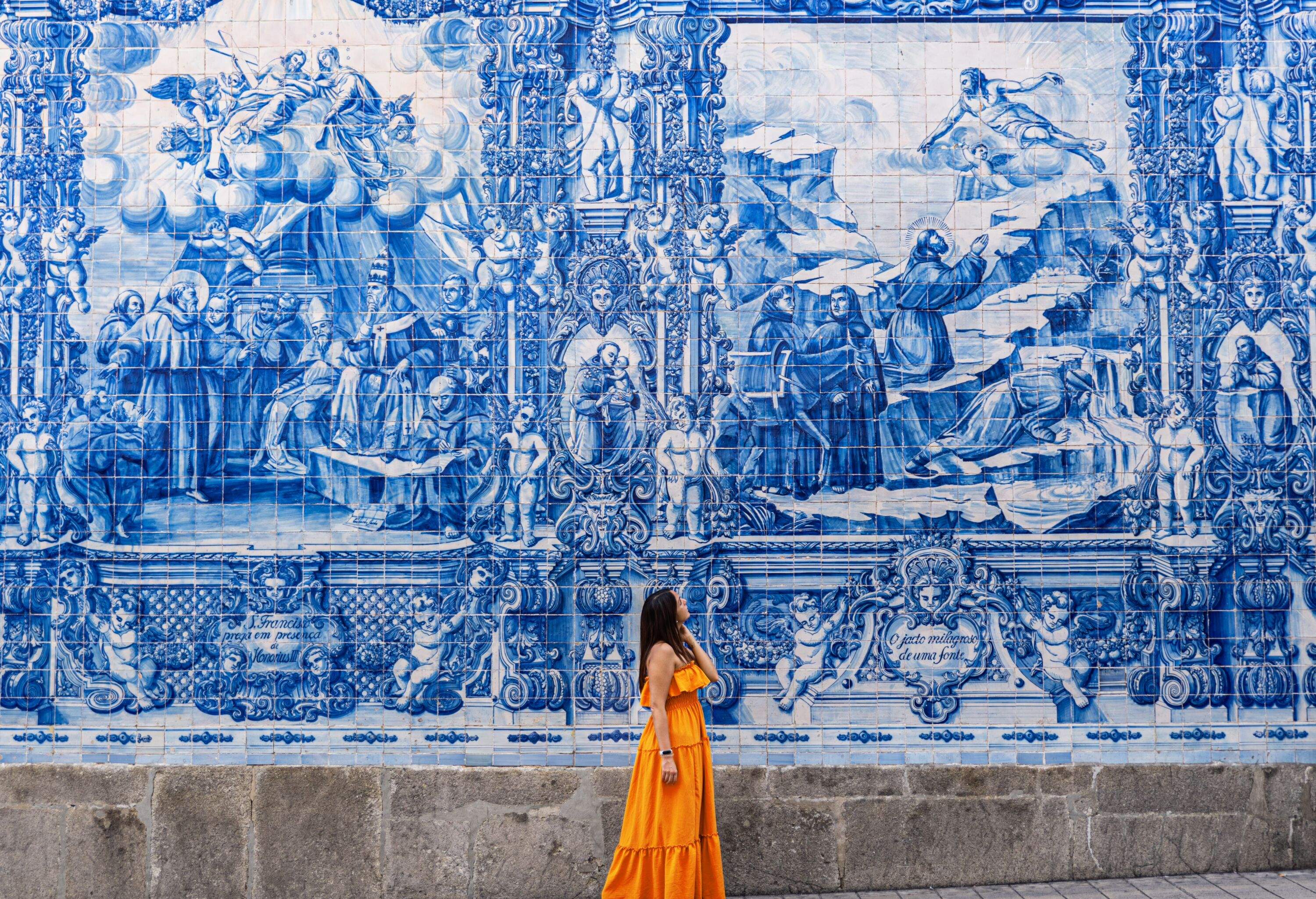 Woman wearing an orange dress standing on a streetwalk wondering at wall tiled with traditional azulejos