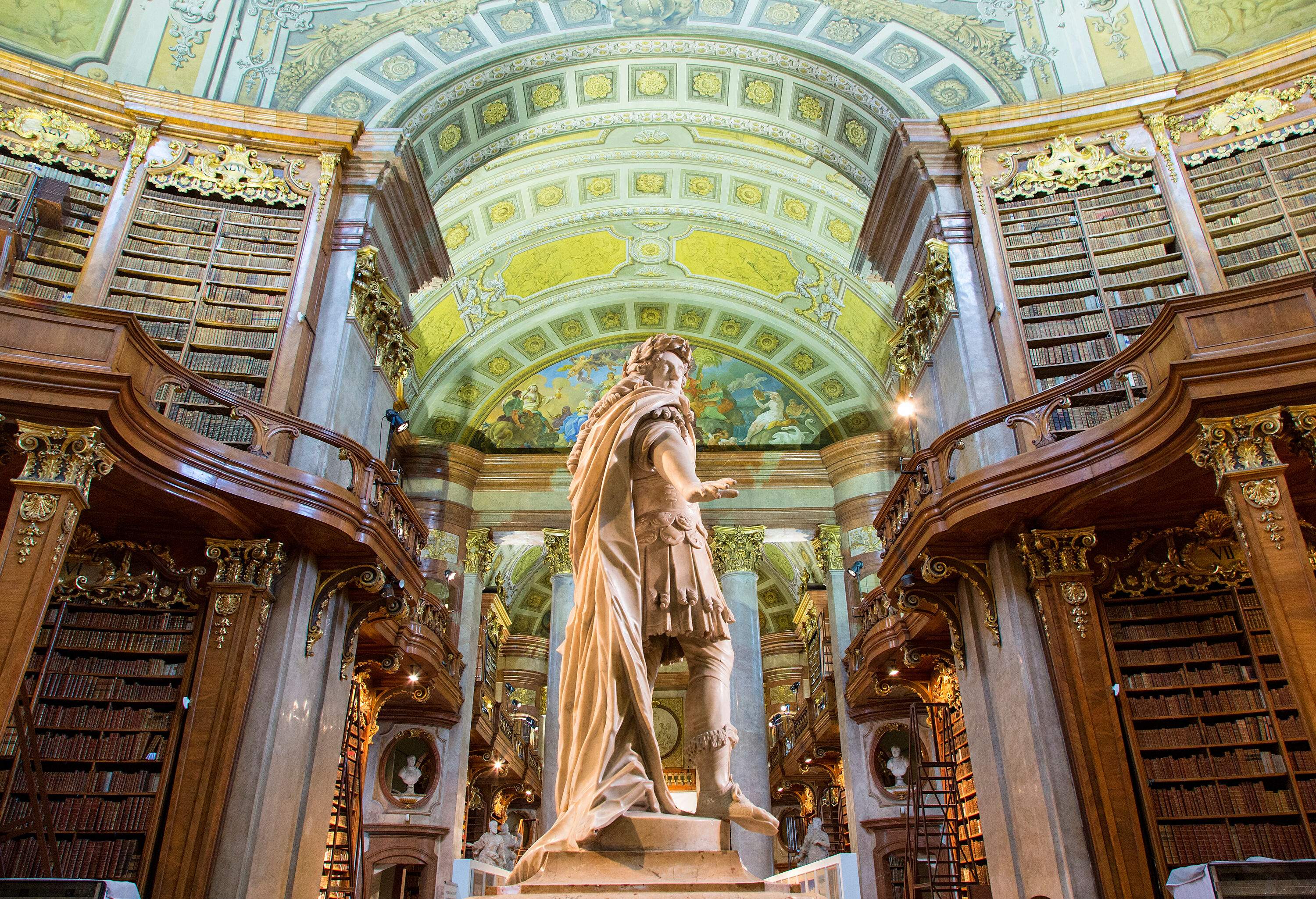 The ceremonial room of the National Library features an imposing statue of Karl VI, with towering bookcases brimming with literary treasures in the background.