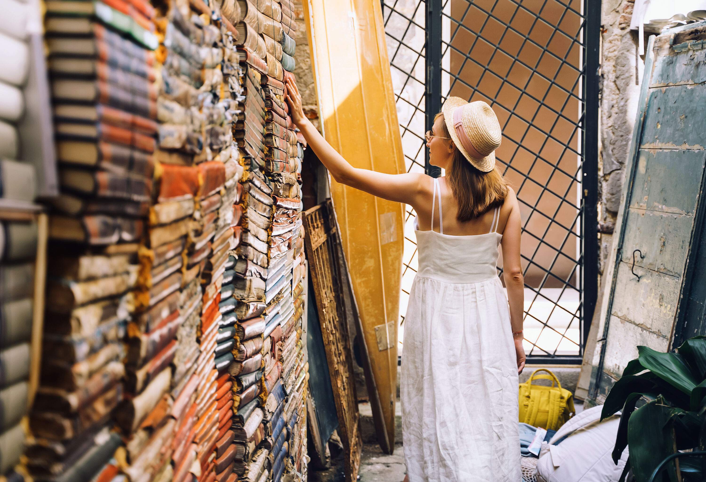 A young woman in a white dress and a hat looking through antique books stacked against the wall.