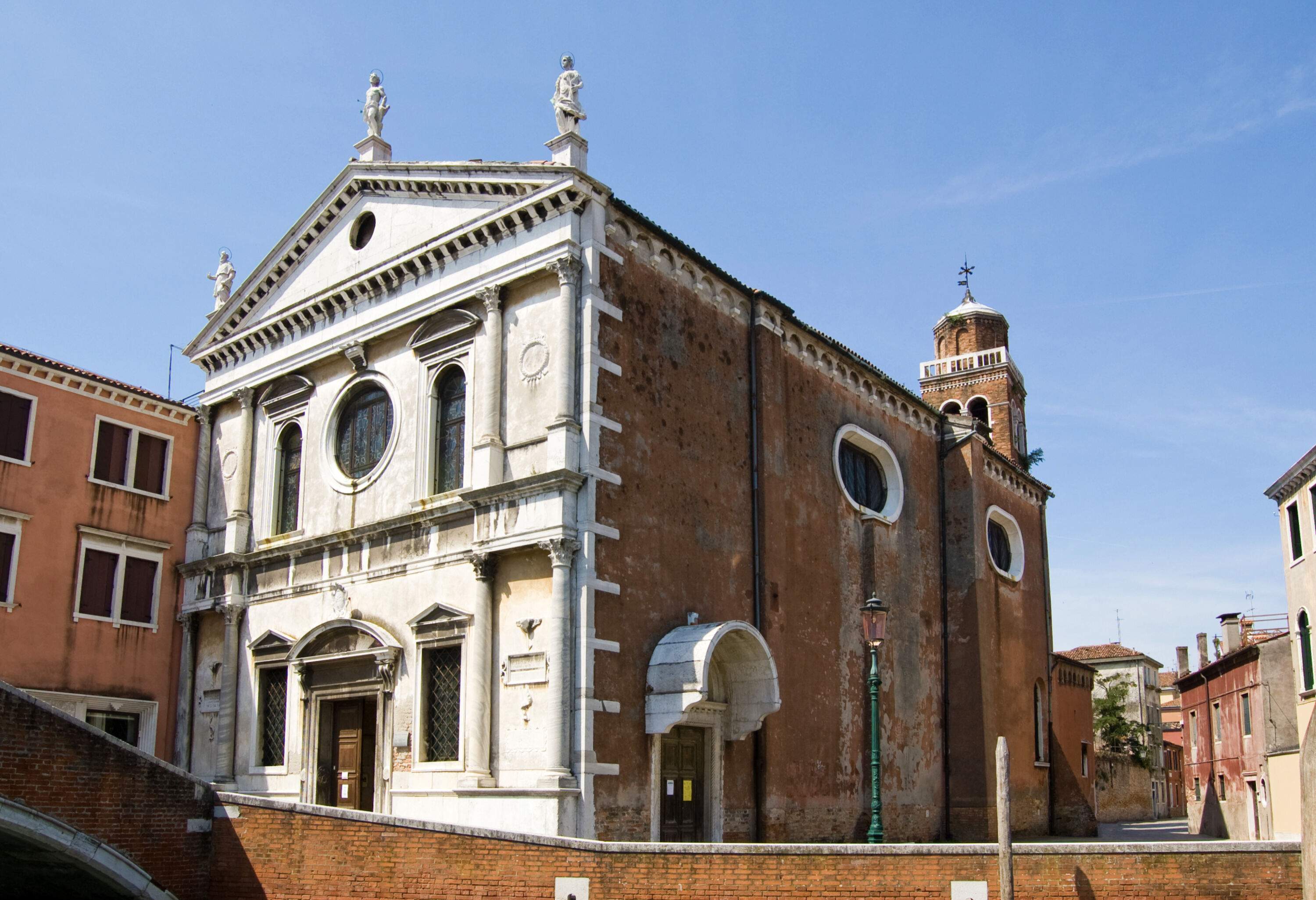 An old Roman Catholic church with a plain white facade in the middle of a cluster of buildings.