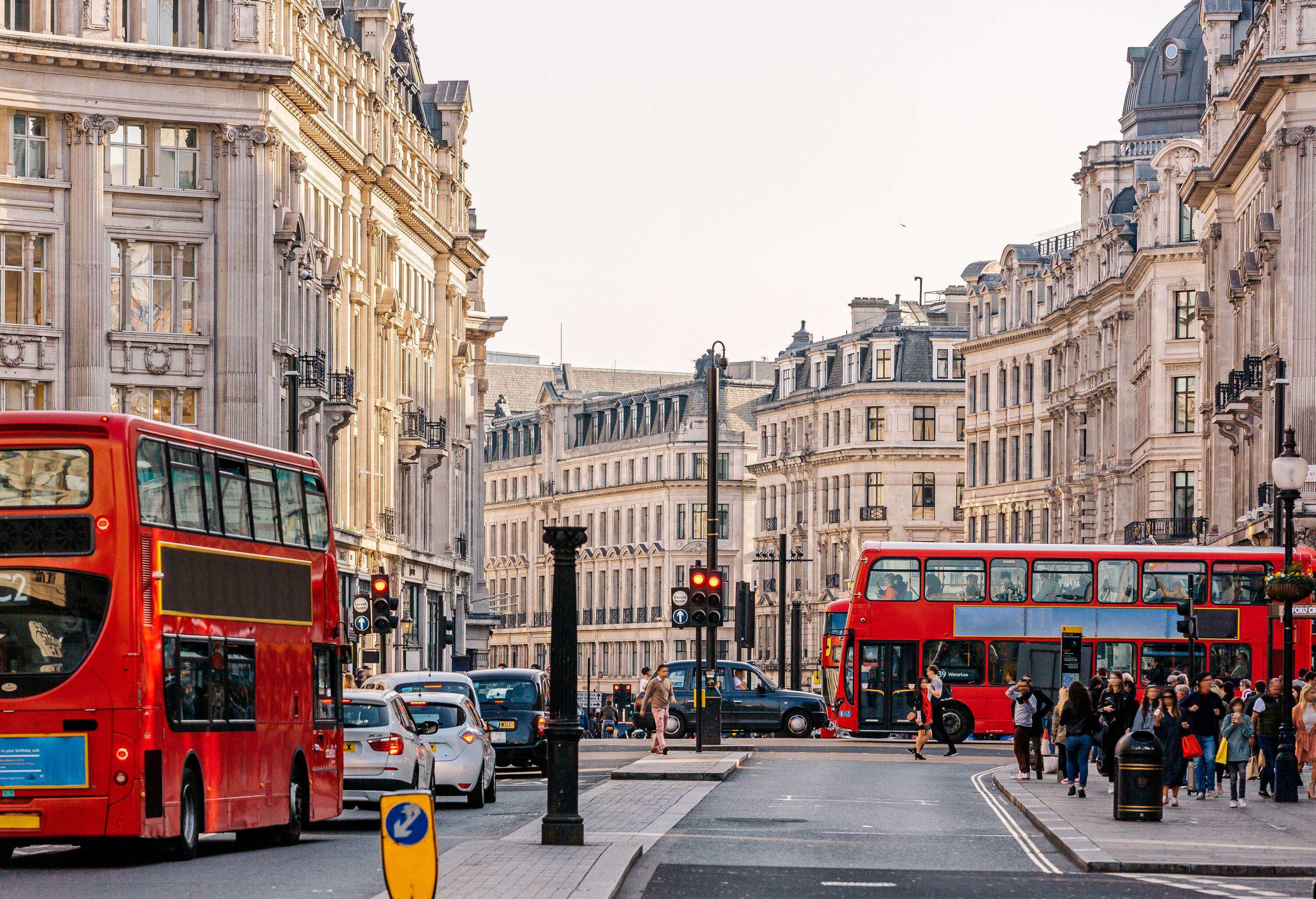 A busy London street with pedestrians walking on the sidewalk and double-decker buses and cars passing by.