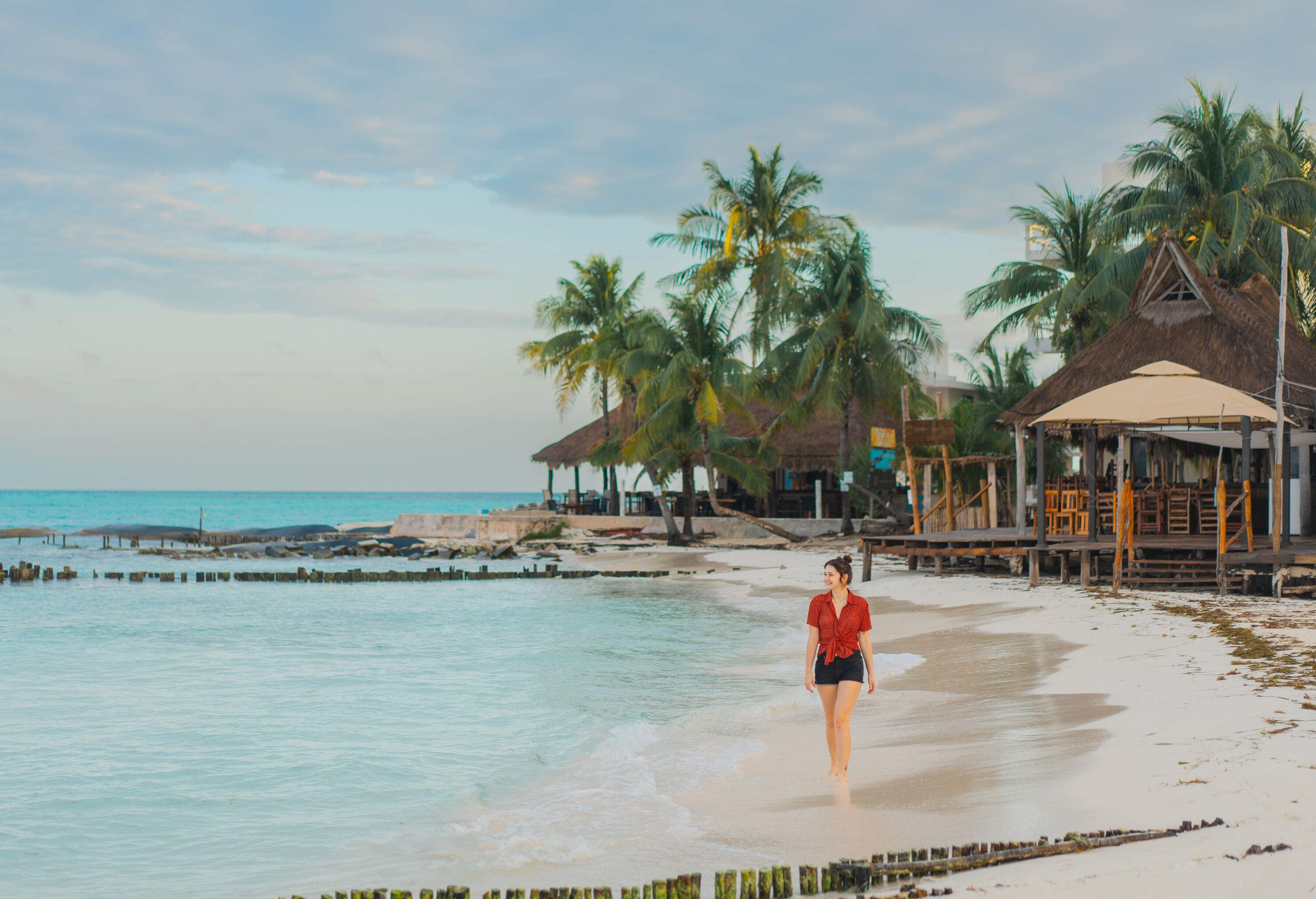 A young Caucasian woman with a smile on her face strolls along the seaside, with traditional thatched huts visible in the background.