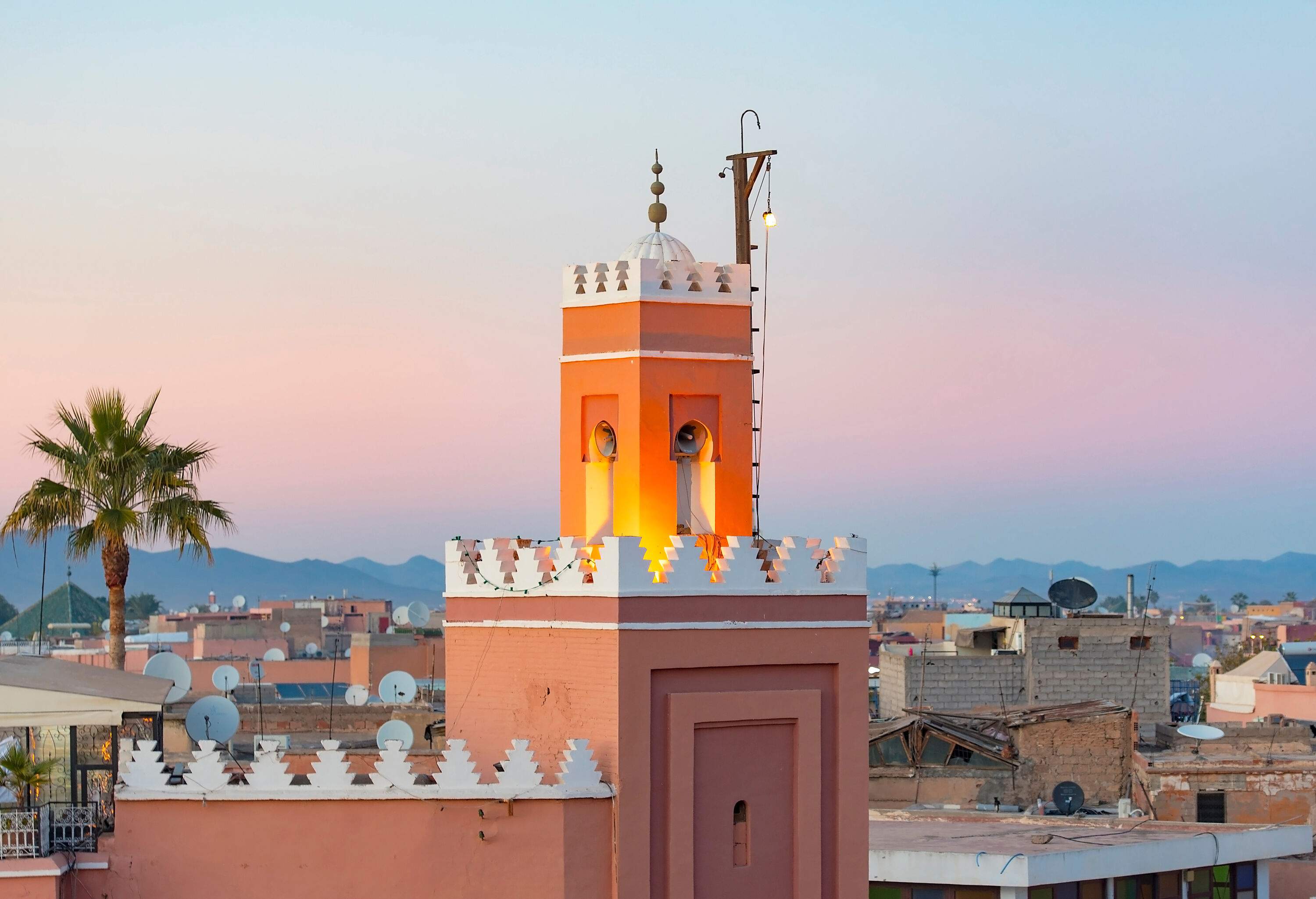 A glowing minaret tower with white decorative battlements in an old city.