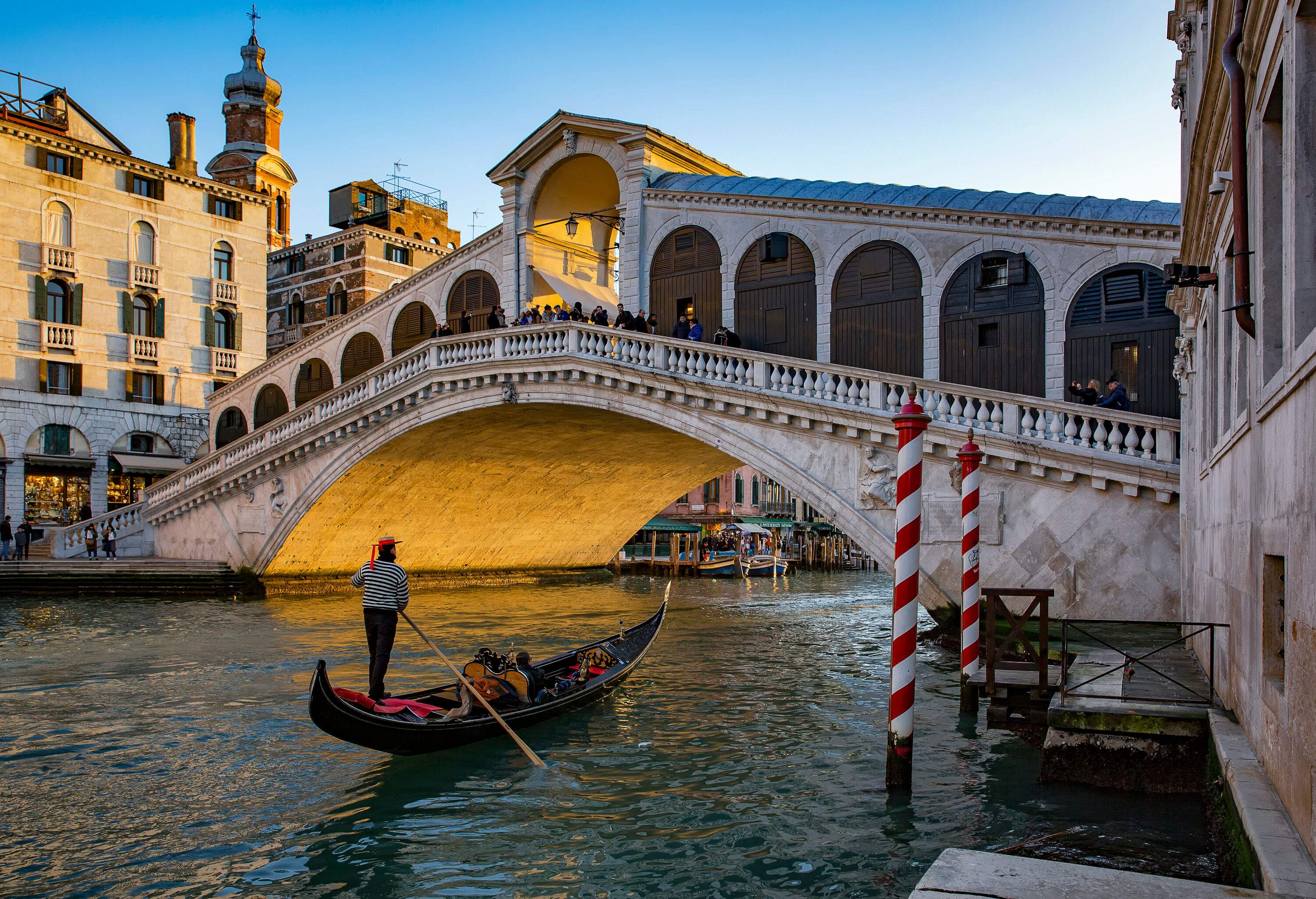 A gondolier on Grand Canal passes the elegant arched Rialto Bridge connecting two classic buildings.