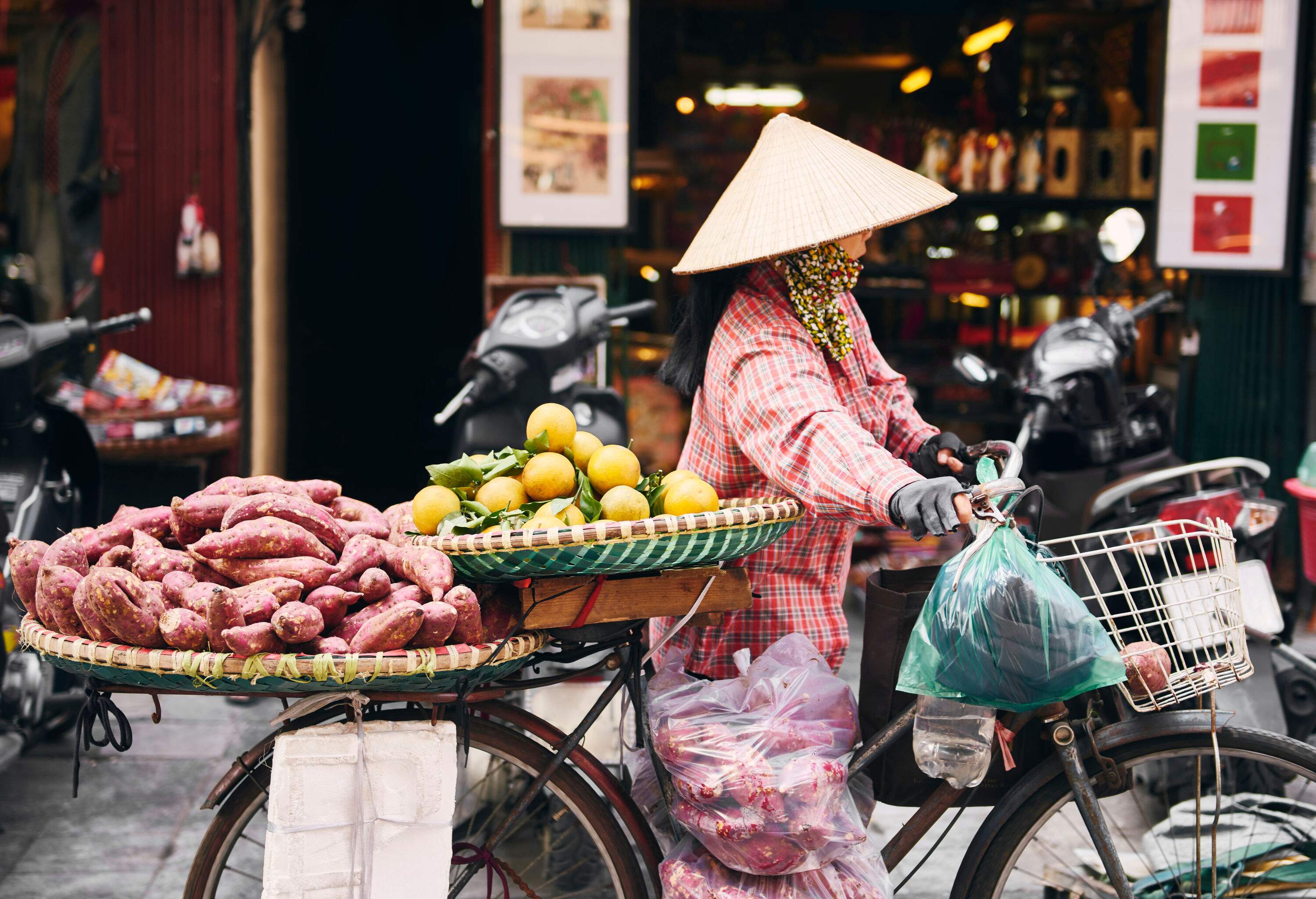 A woman wearing a conical hat walking her bike loaded with round straw trays of sweet potatoes and oranges.