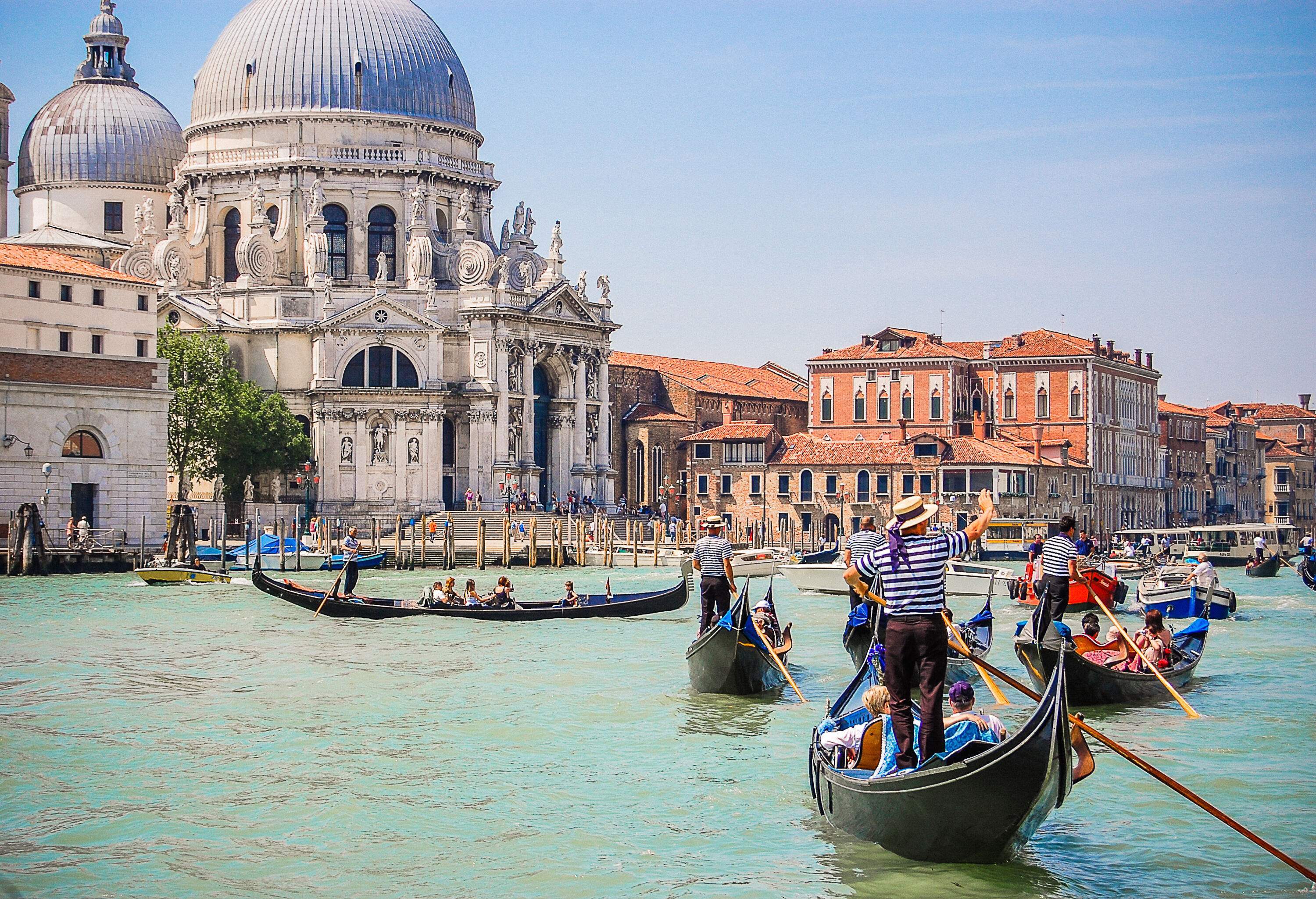 Tourists ride gondolas and other various boats on the Grand Canal with a view of an iconic dome church and other colourful buildings.