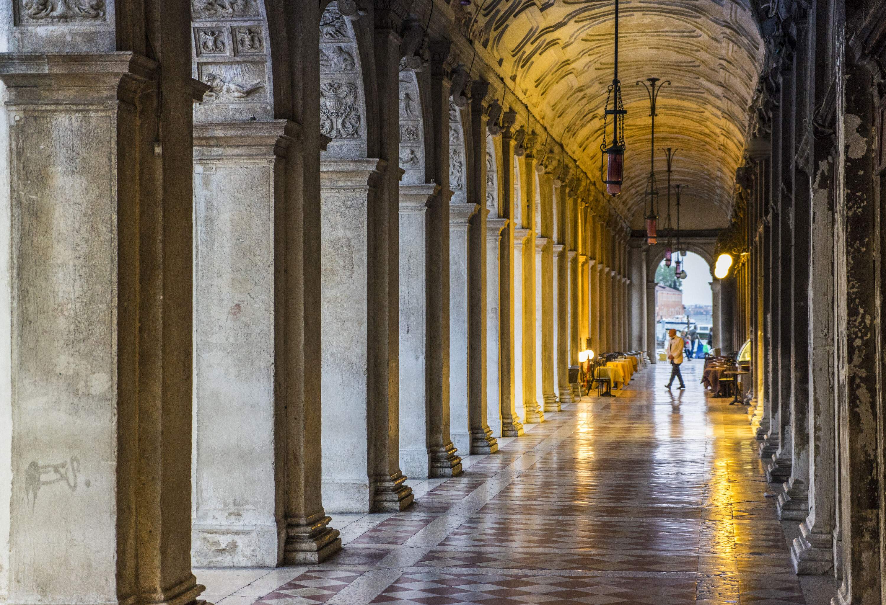 A person walks across a pillared hallway with bright lights and outdoor dining area.