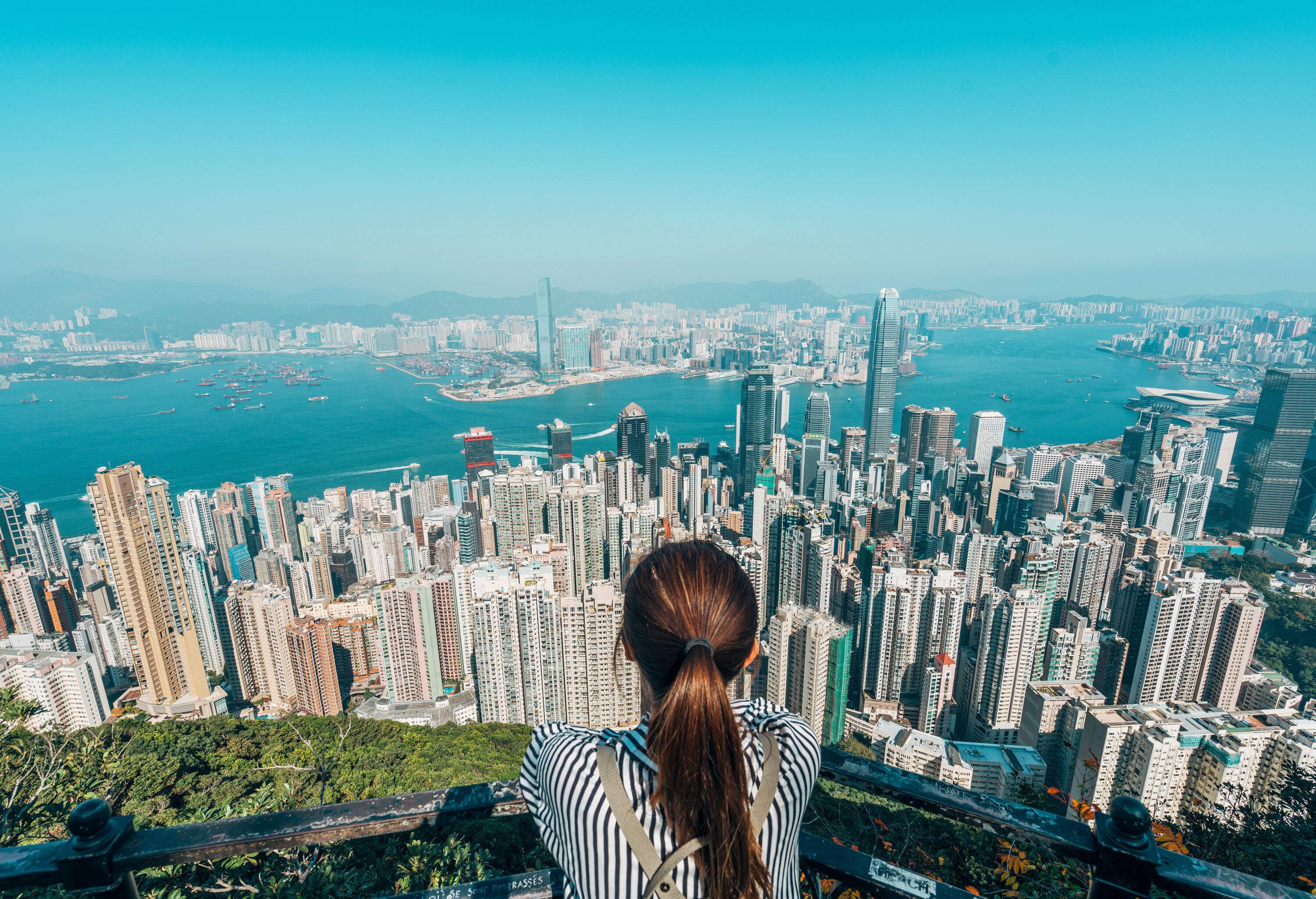 A woman leans on an iron railing on a view deck with a beautiful cityscape on the coast bisected by a long channel flowing amidst the tall buildings.