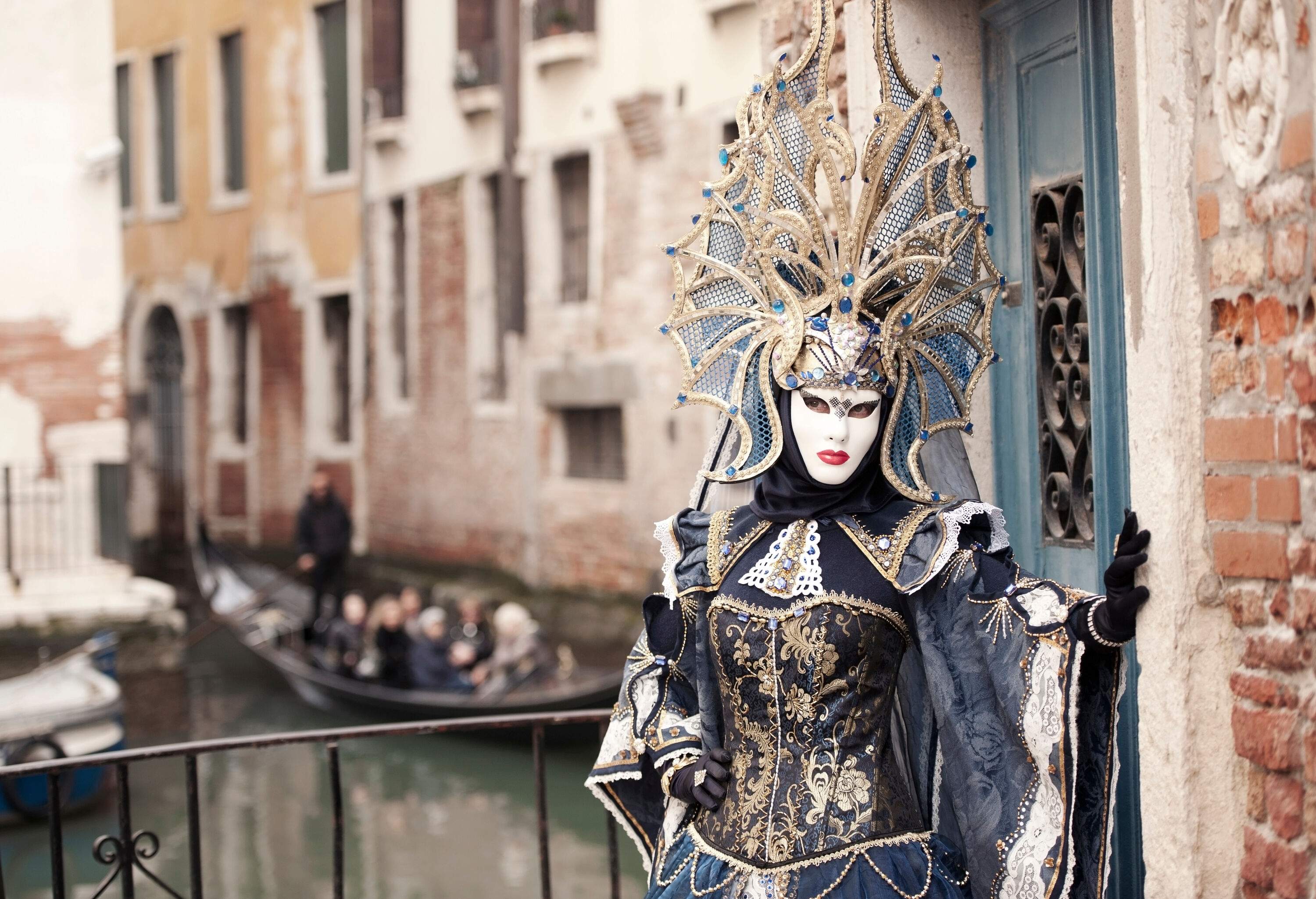 A colourful and attractive figure, dressed in a glamorous carnival costume and adorned with an ornate headpiece, poses in front of a canal.