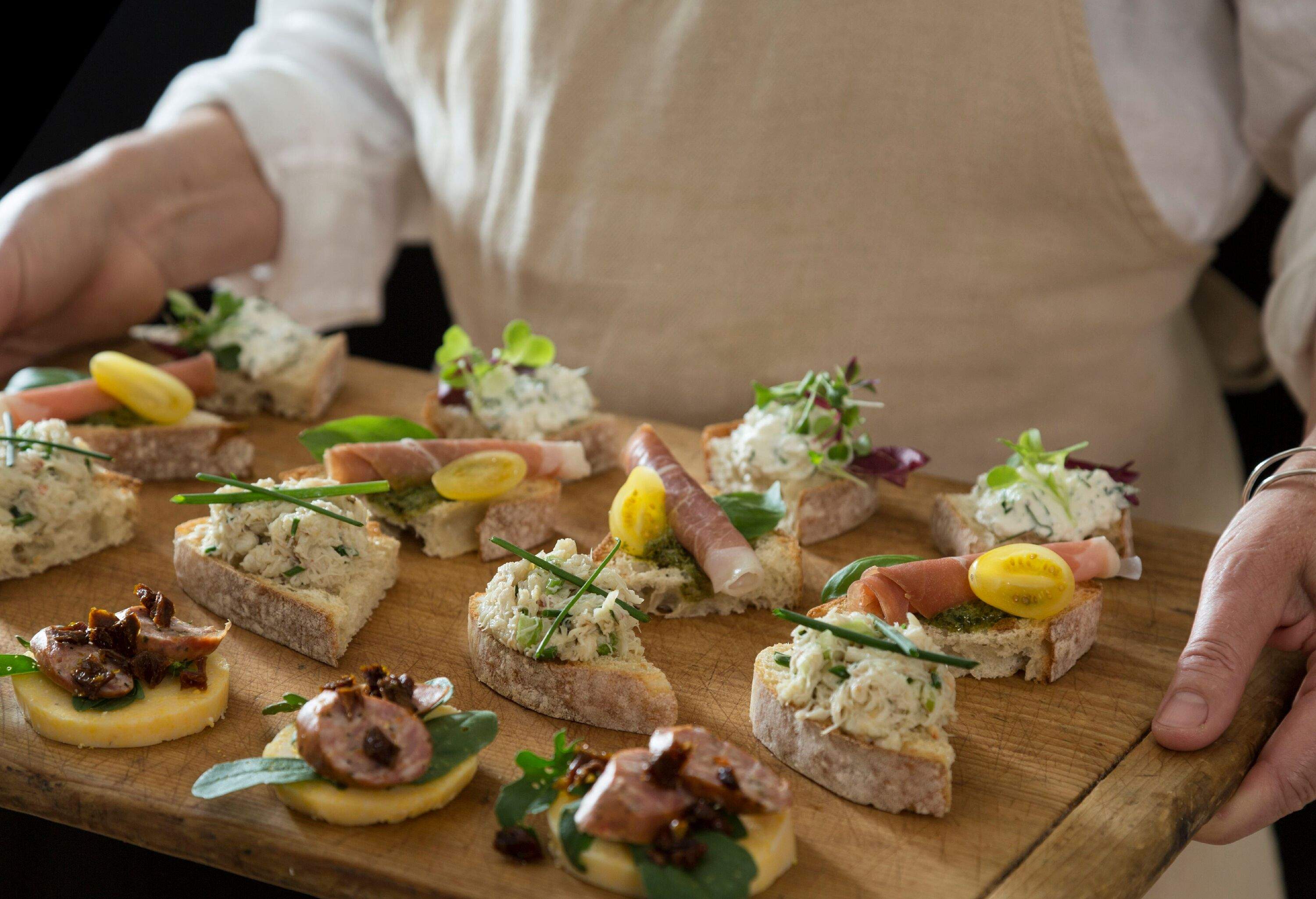 A hospitable individual wearing an apron proudly presents an assortment of delectable cicchetti on a rustic wooden board.