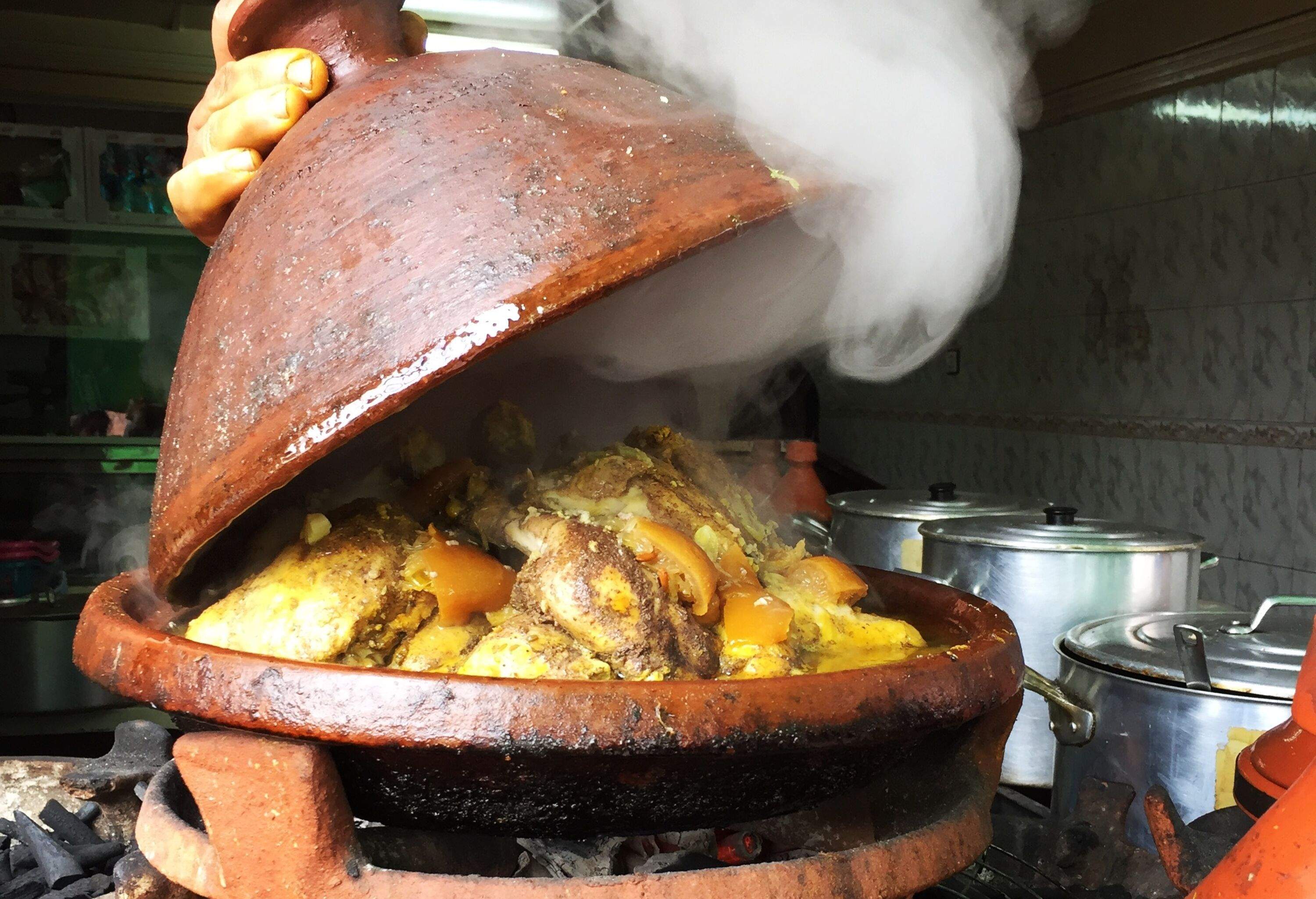 A hand opens the lid of a clay pot with a steaming and savoury meat stew.