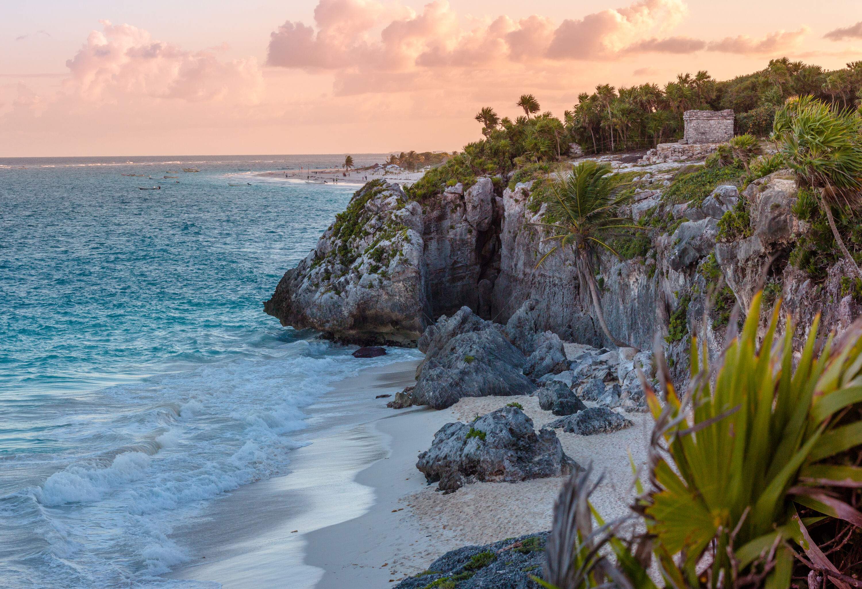 The majestic Tulum ruins, perched atop a coastline cliff, are bathed in the warm glow of the sunset, with waves crashing against the shore and lush, verdant surroundings framing the idyllic beach below.