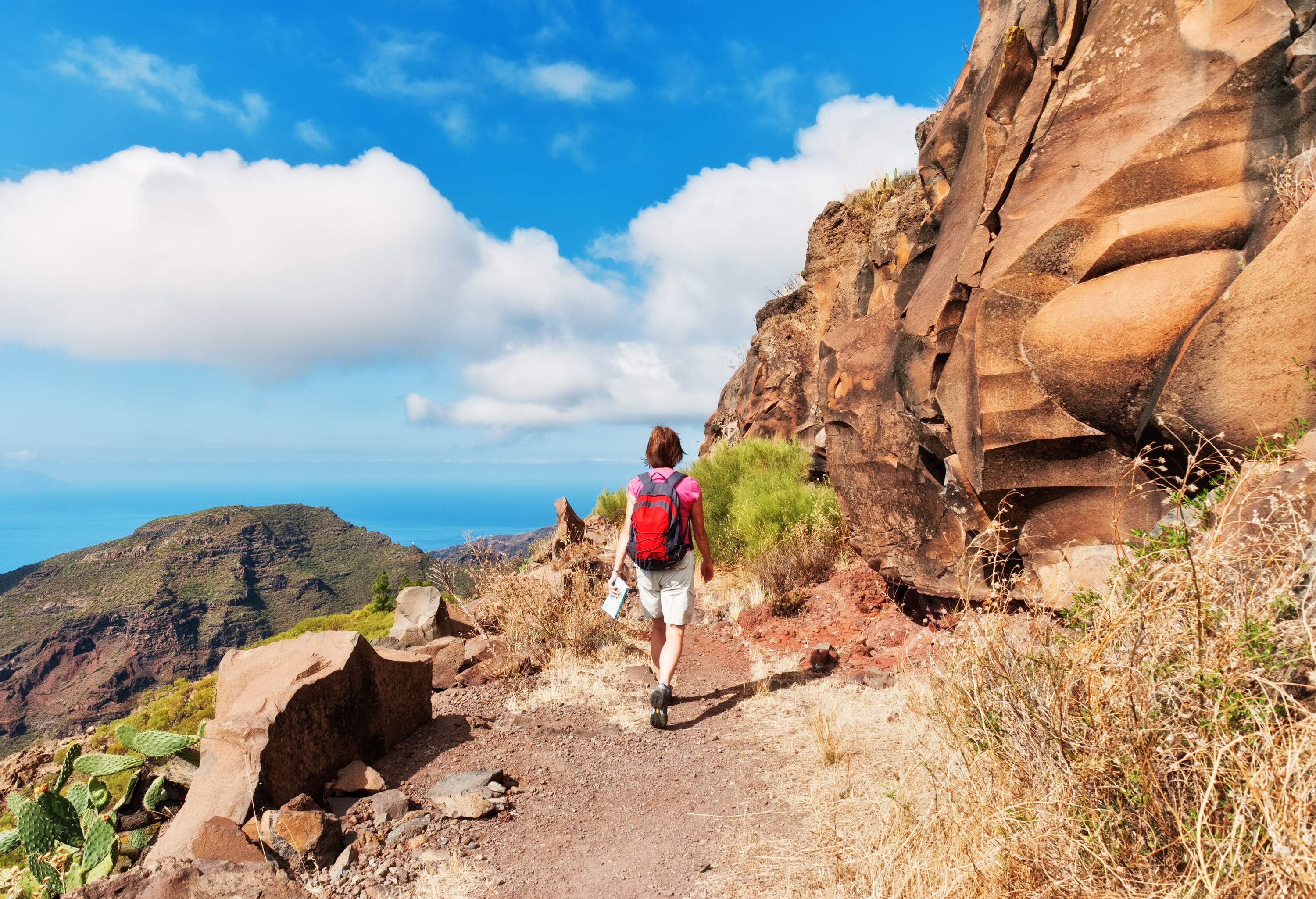 A hiker carrying a red bag walks along a mountainside trail that features unusual rock walls.