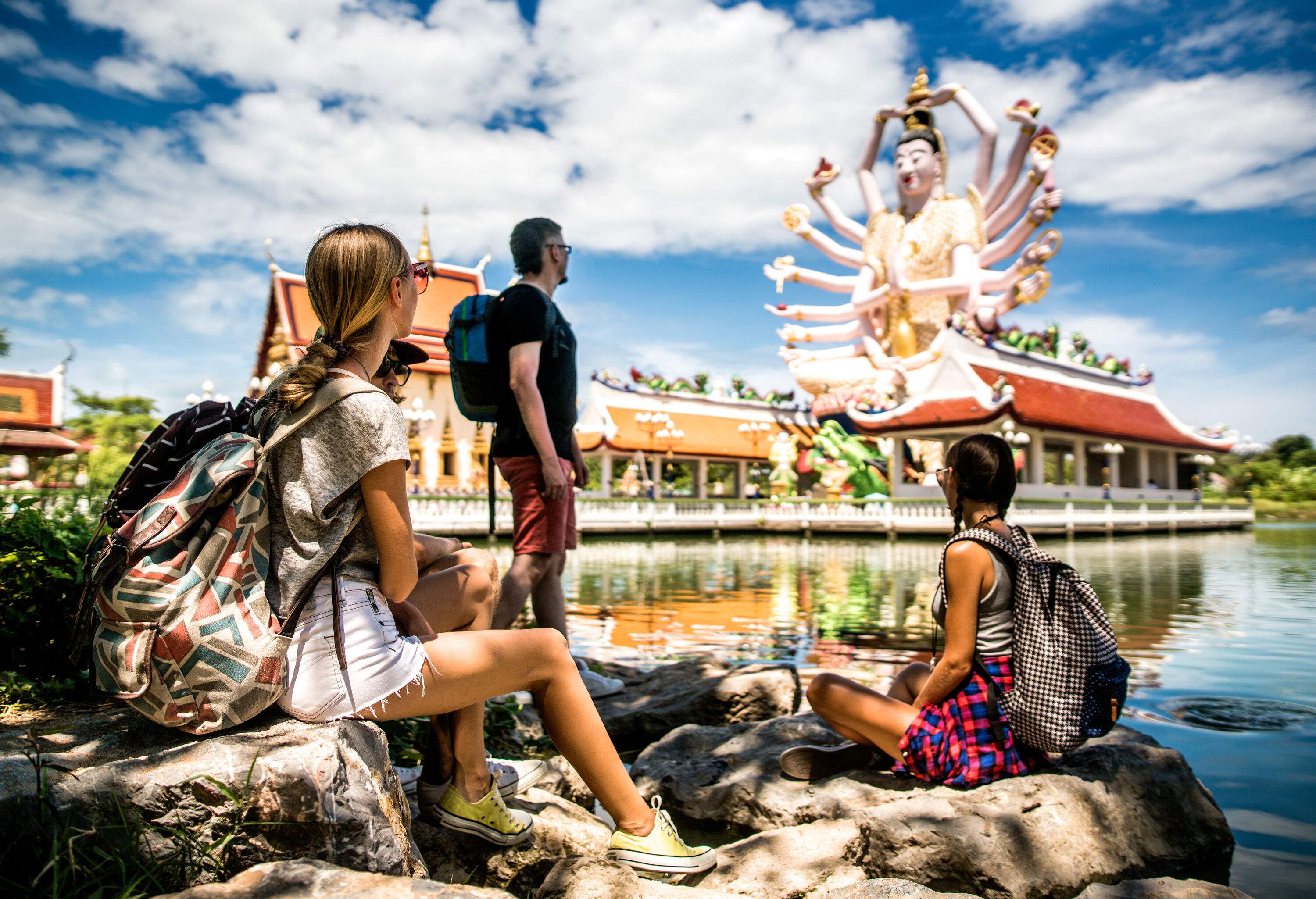 A group of friends on tour alongside the lake look at the sixteen-armed statue atop an open temple.