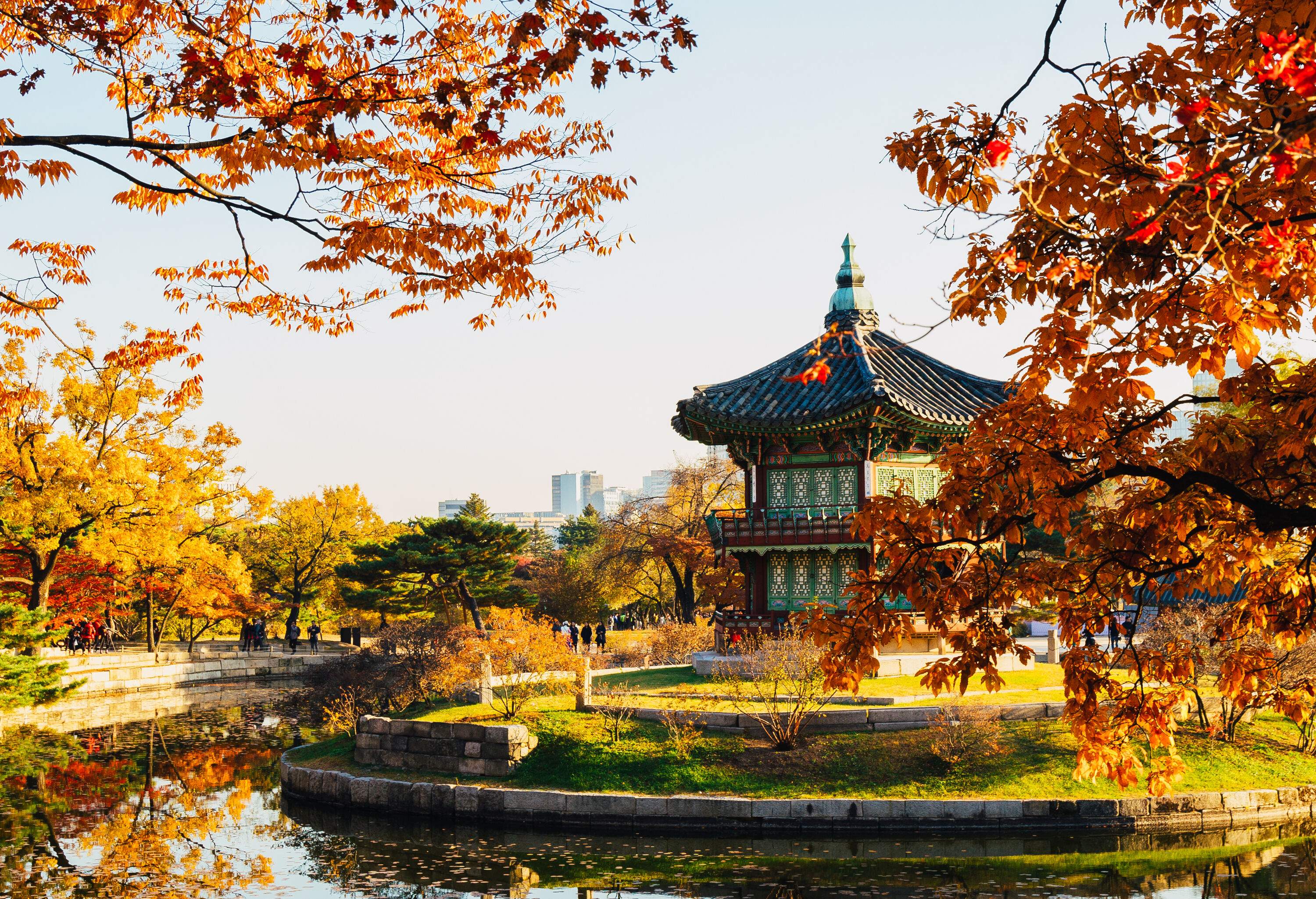 Perched on a small island in the middle of a square pond is a small two-story hexagonal historic Korean pavilion surrounded by trees in vibrant autumn colours.