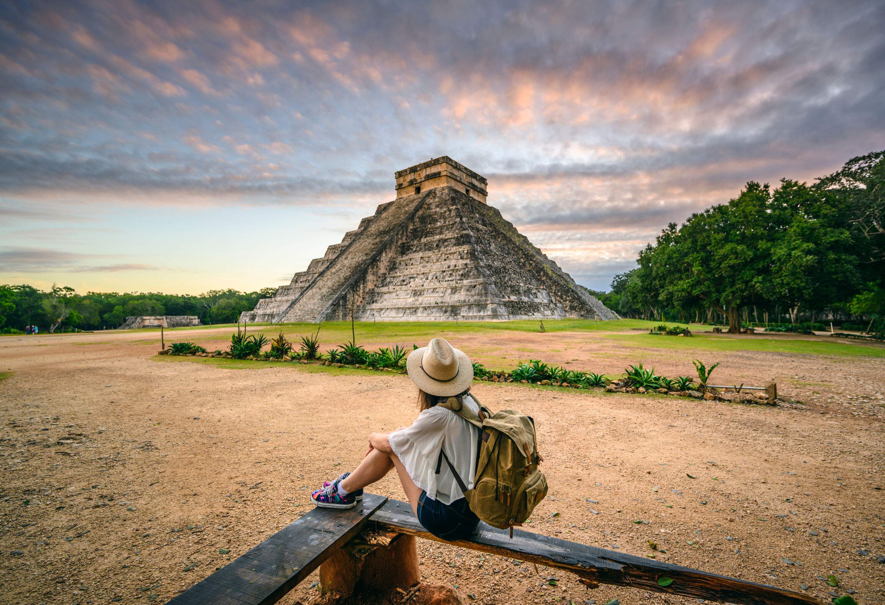 A young lady sits on a rugged wooden bench looking at the ancient temple pyramid surrounded by lush trees.