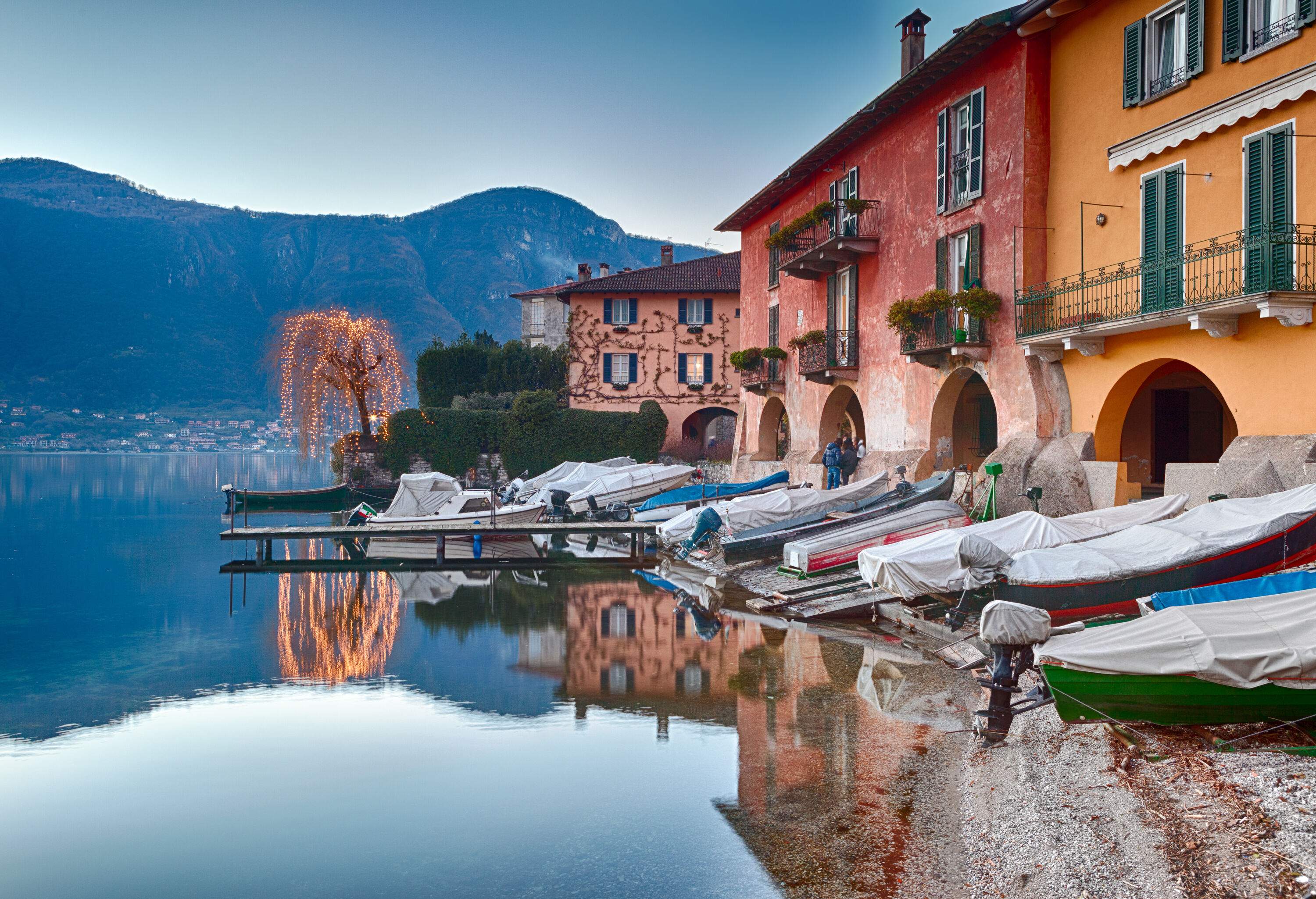 A row of covered boats docked alongside the coastal buildings on a lakeshore.