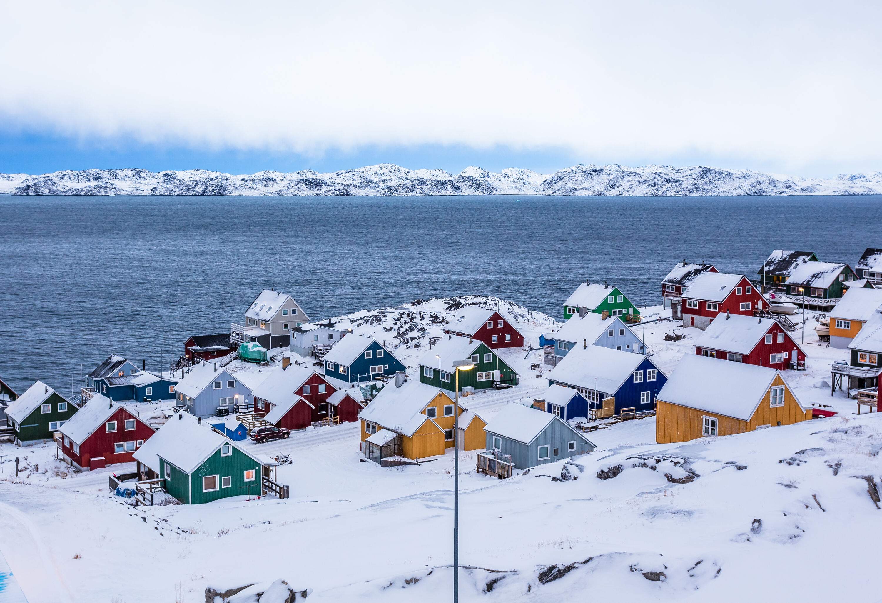 Colourful Inuit houses nestled among rocky terrain and snow-covered surroundings, situated by a fjord in a suburban area.