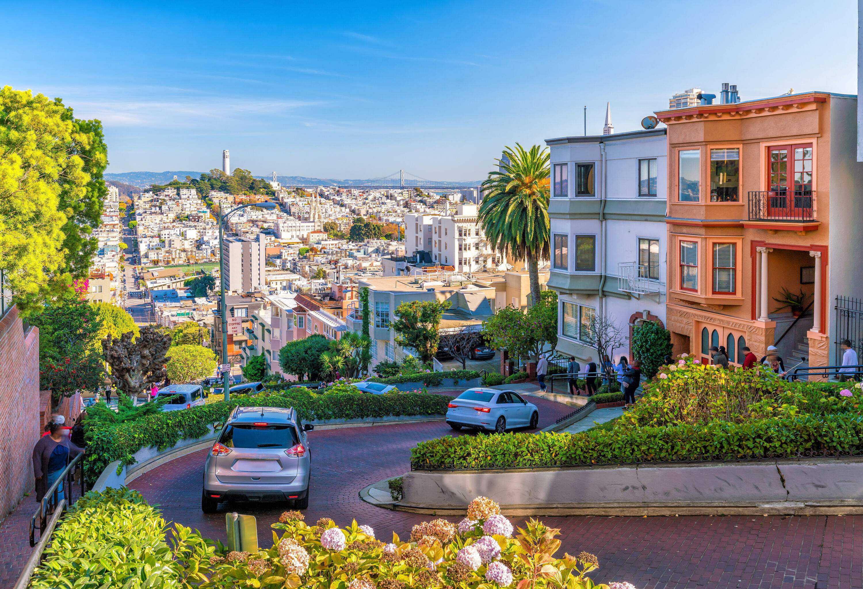 A couple of cars descending a sloping street with colourful houses and a view of buildings across the valley.