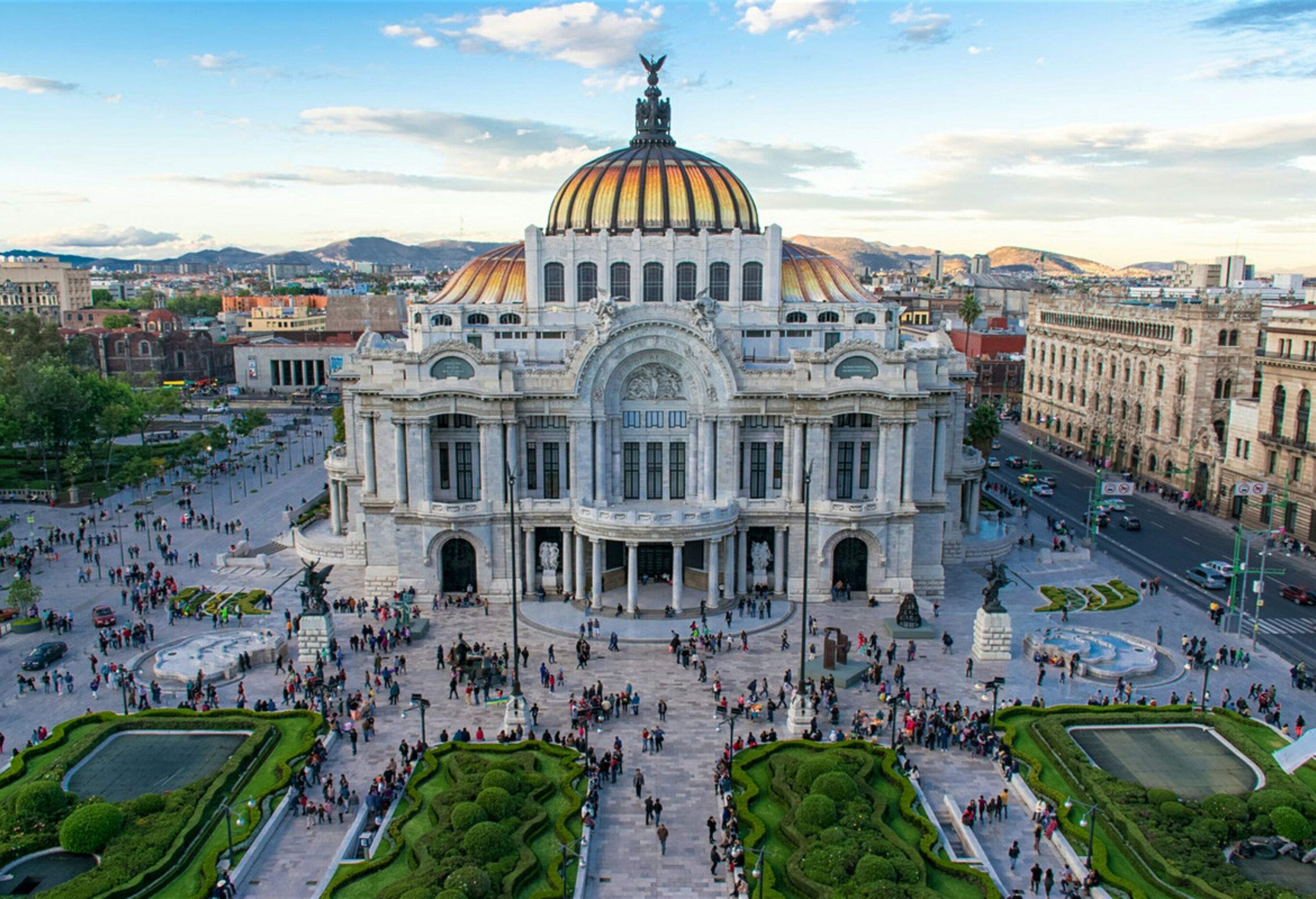 The stunning Palacio de Bellas Artes boasts an elegant white marble facade, intricate metalwork, and vibrant stained-glass windows, combining Art Nouveau and Art Deco styles, while its beautiful gardens and people strolling about create a peaceful and charming atmosphere around this cultural gem.