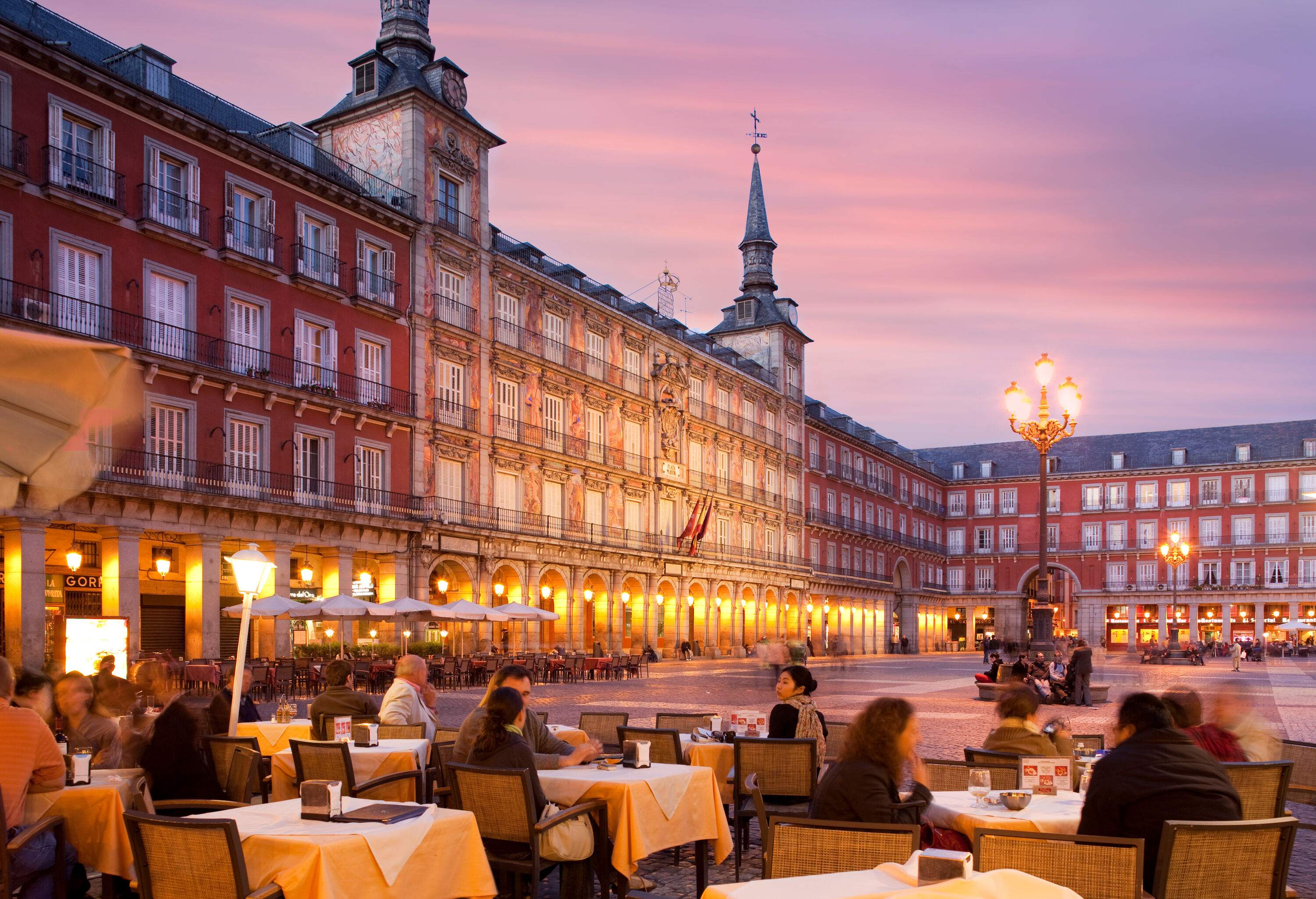 Customers dine alfresco at a plaza restaurant surrounded by lit buildings.