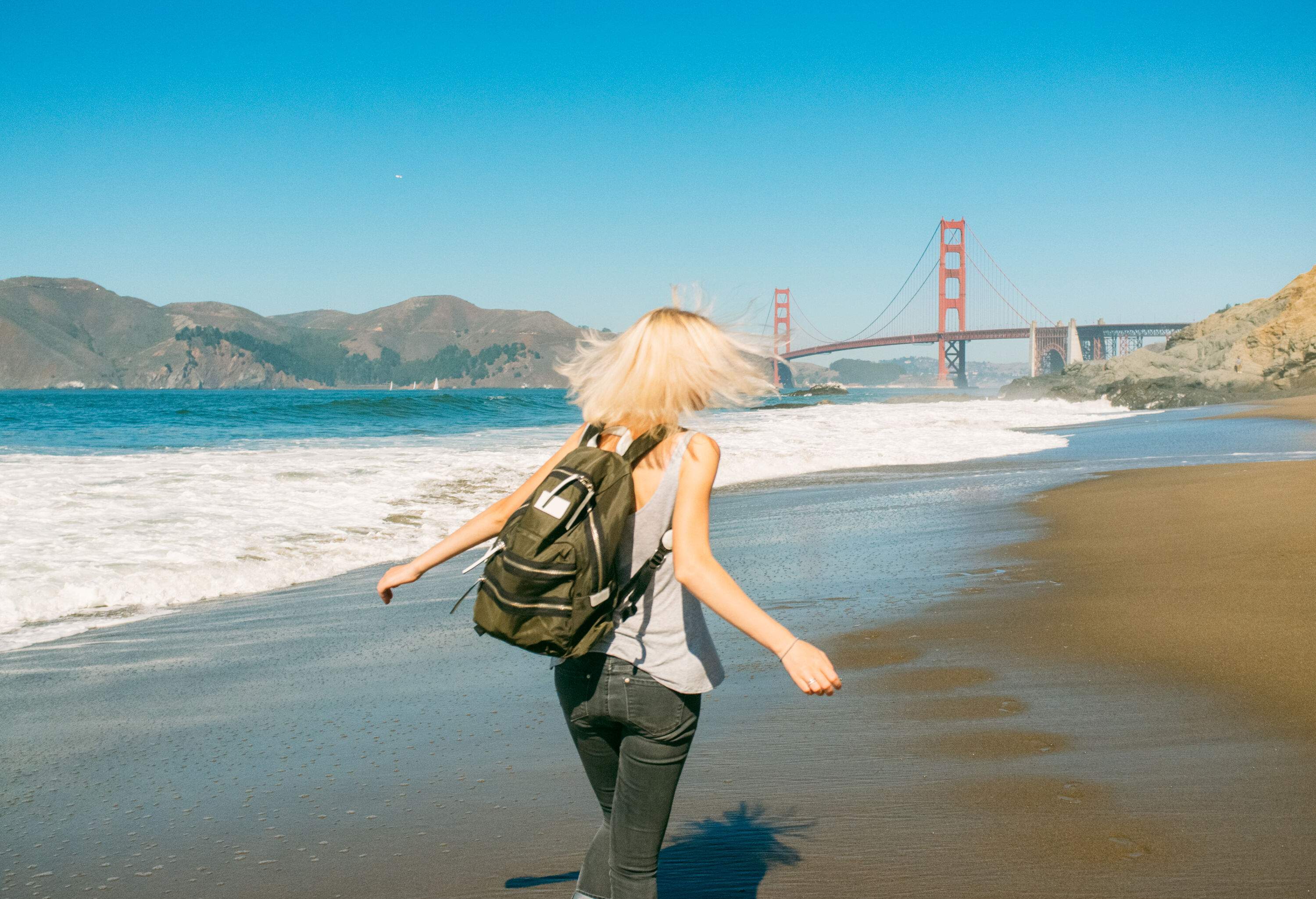 A sun-kissed blonde individual strolls along the beach, donning a backpack, while the majestic Golden Gate Bridge stands proudly in the distance.