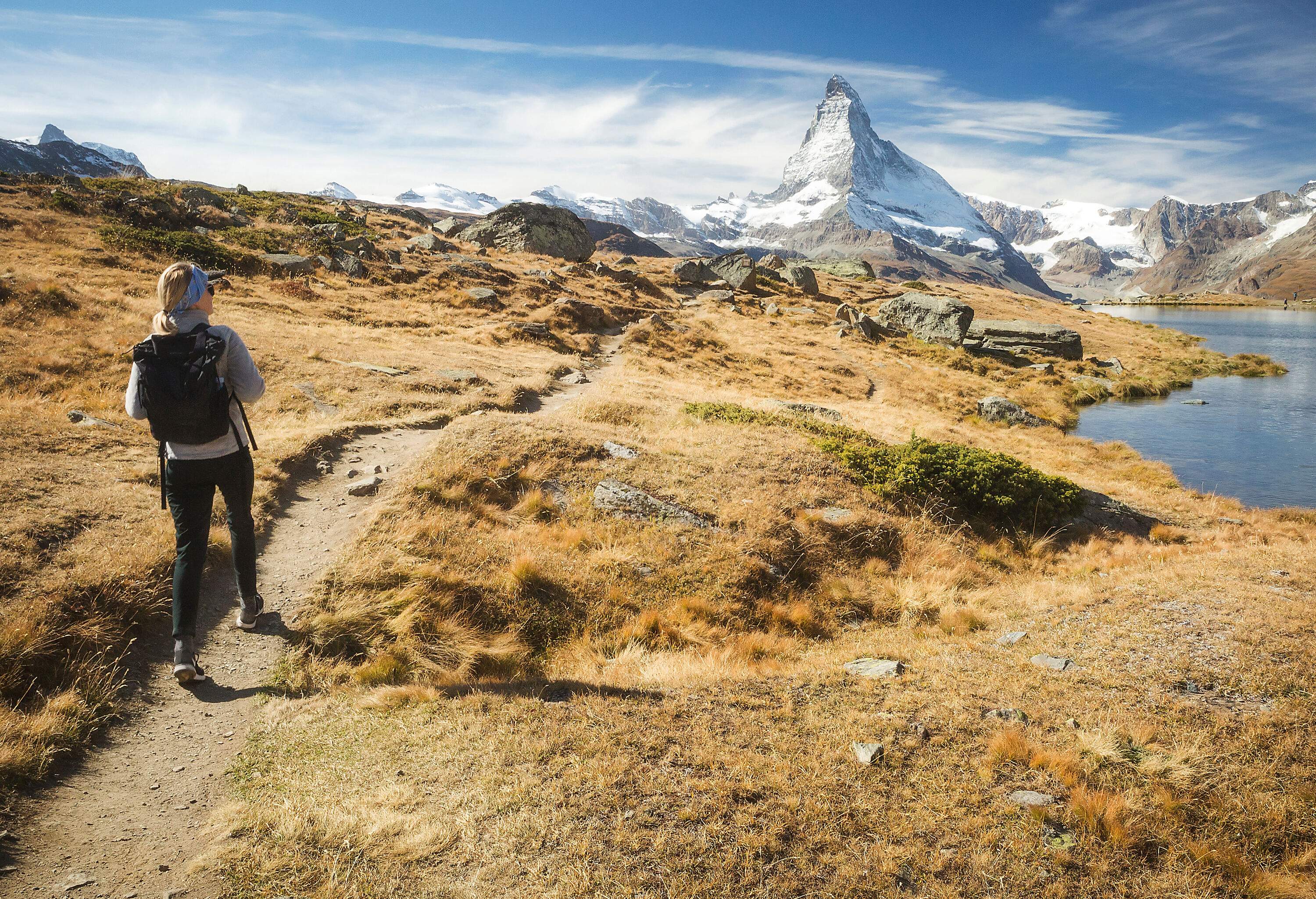 A hiker with a black backpack walks along a lakeside path overlooking the snow-dusted rocky mountains.