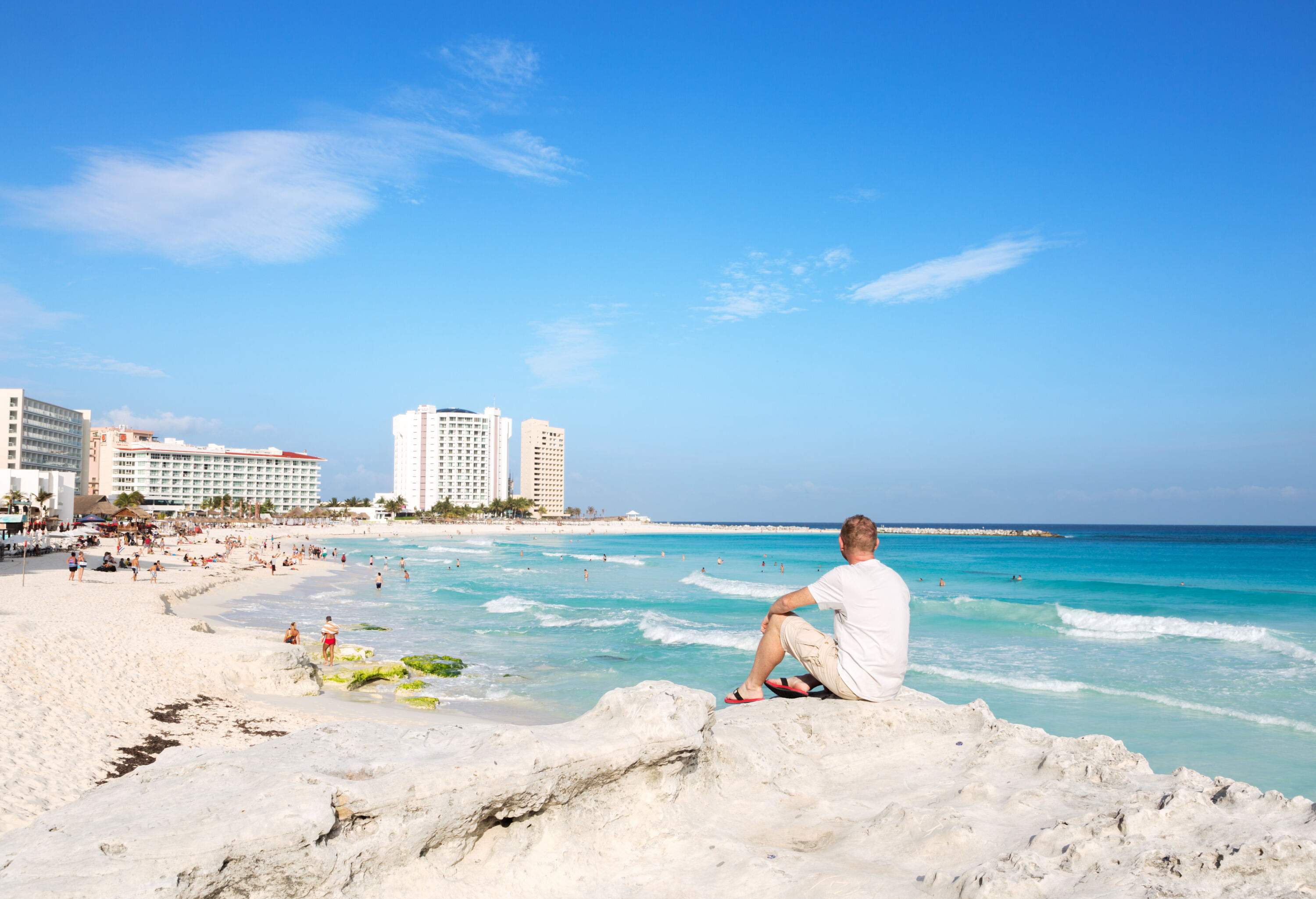 A person sits contemplatively by a rock, gazing towards the beach ahead, where beachgoers wander about, while buildings in the distance create a backdrop of coastal charm.