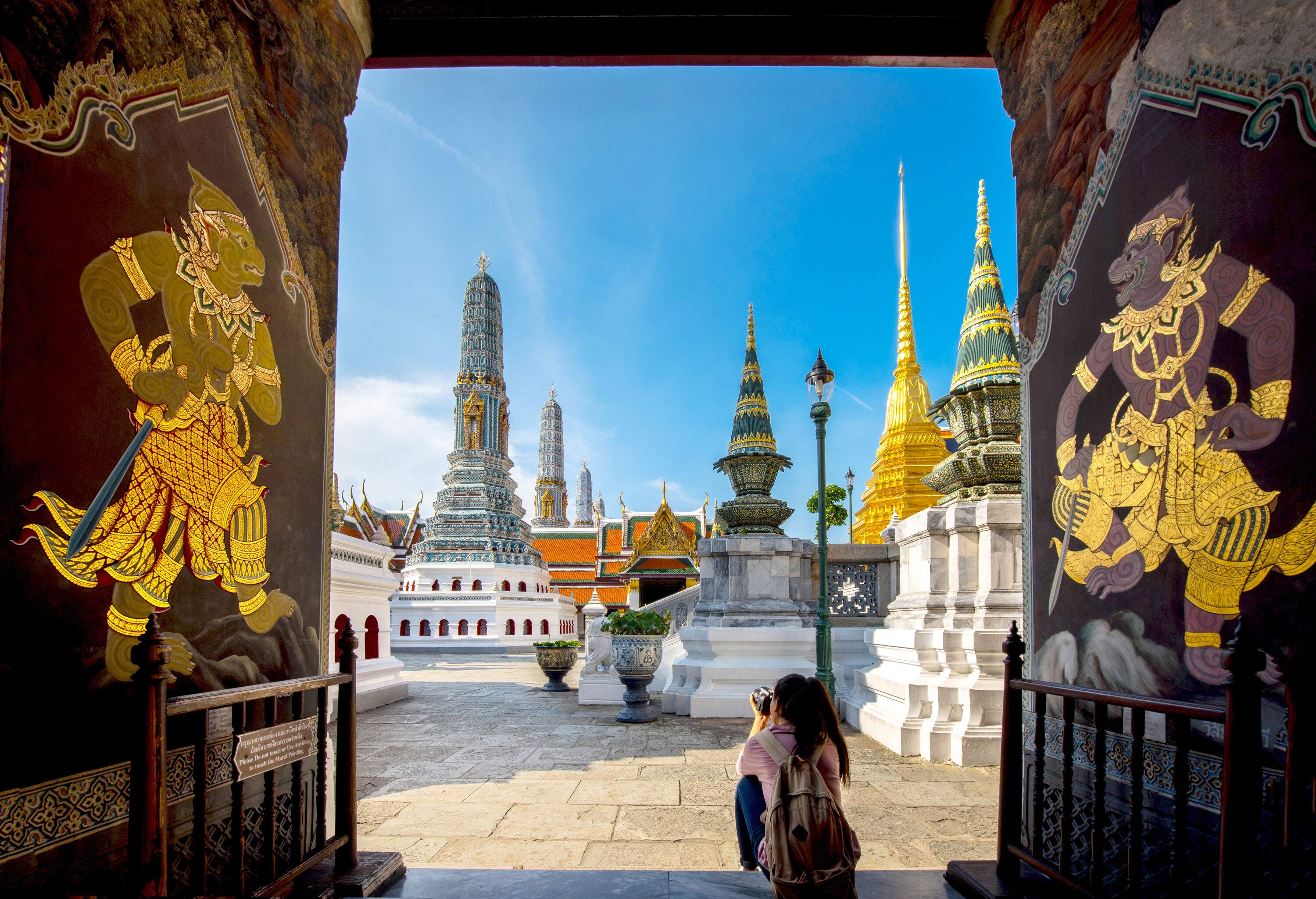 A tourist sits at the door of a temple decorated with Ramanaya images, taking pictures of the pagoda-like structure of the Grand Palace in Bangkok, Thailand.