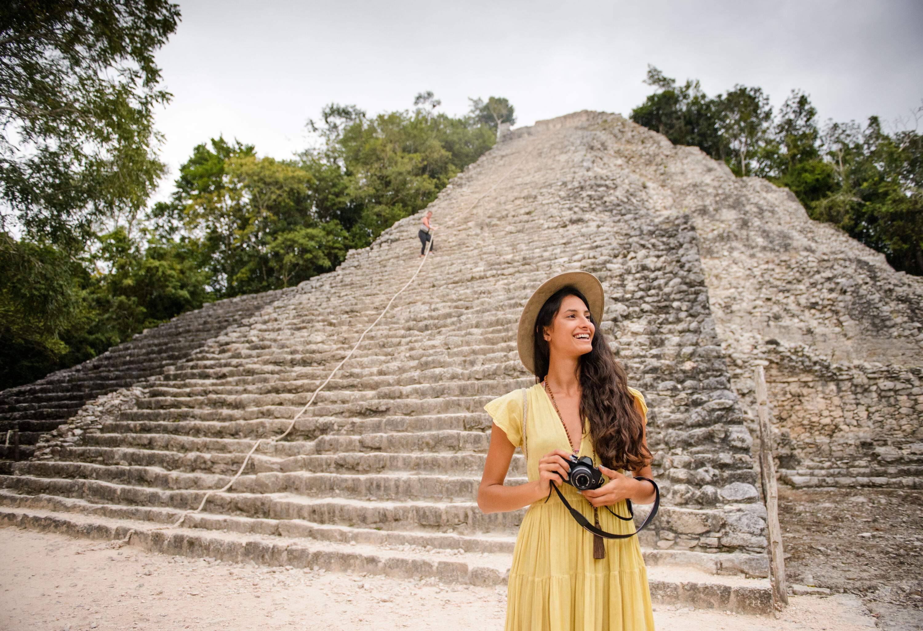 A happy woman in a yellow dress holds a camera and stands against a historic pyramid surrounded by tall green trees.